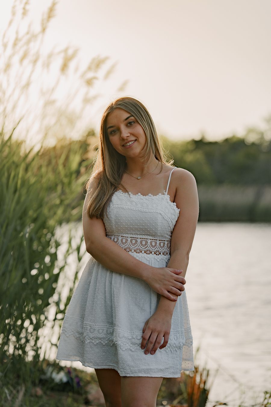 Young woman in white lace sundress smiling by a sunlit lake with tall grass.