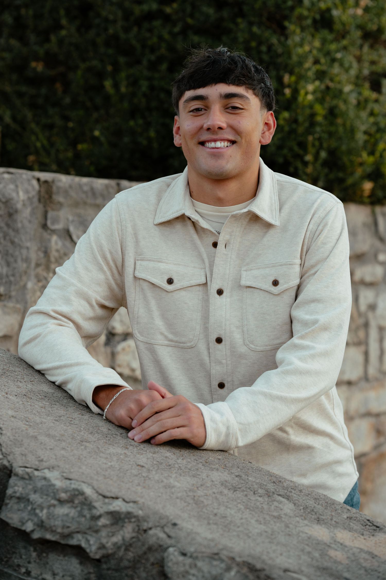 Person leaning on a stone wall wearing a light-colored button-up jacket in an outdoor setting.