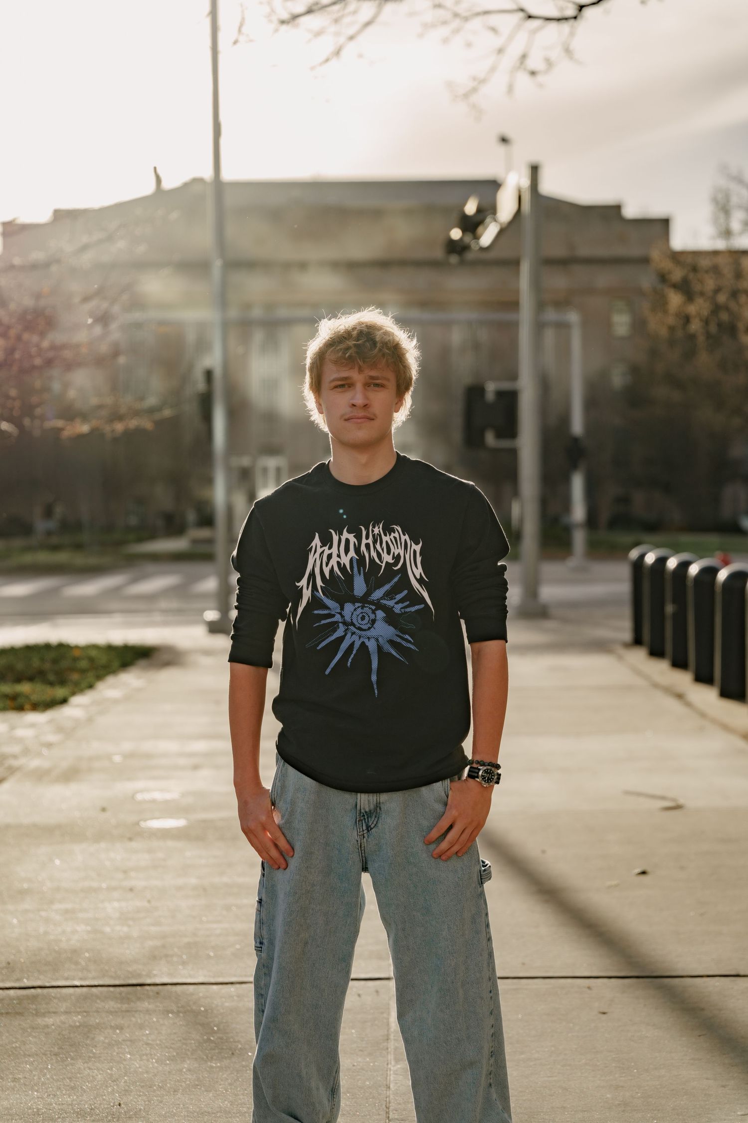 Young man in black graphic tee and jeans standing on urban sidewalk in natural sunlight.
