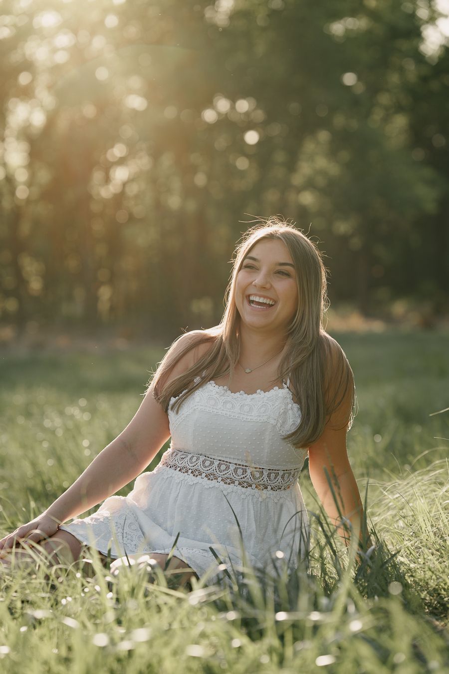 Young woman in white dress sitting in sunlit meadow, laughing joyfully with golden hour light filtering through trees.