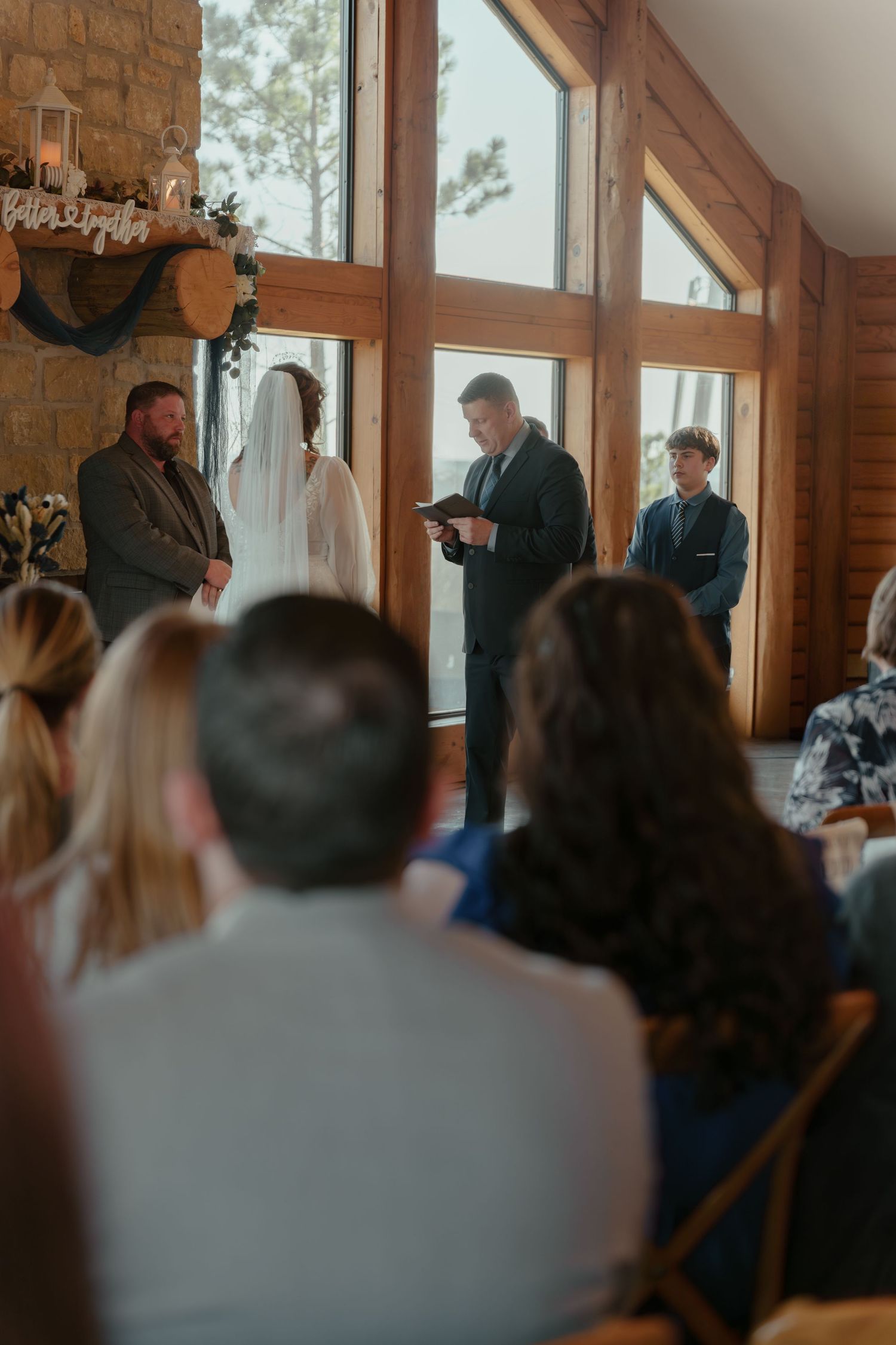 Indoor wedding ceremony in a rustic wood chapel with bride, groom, and officiant before large windows.