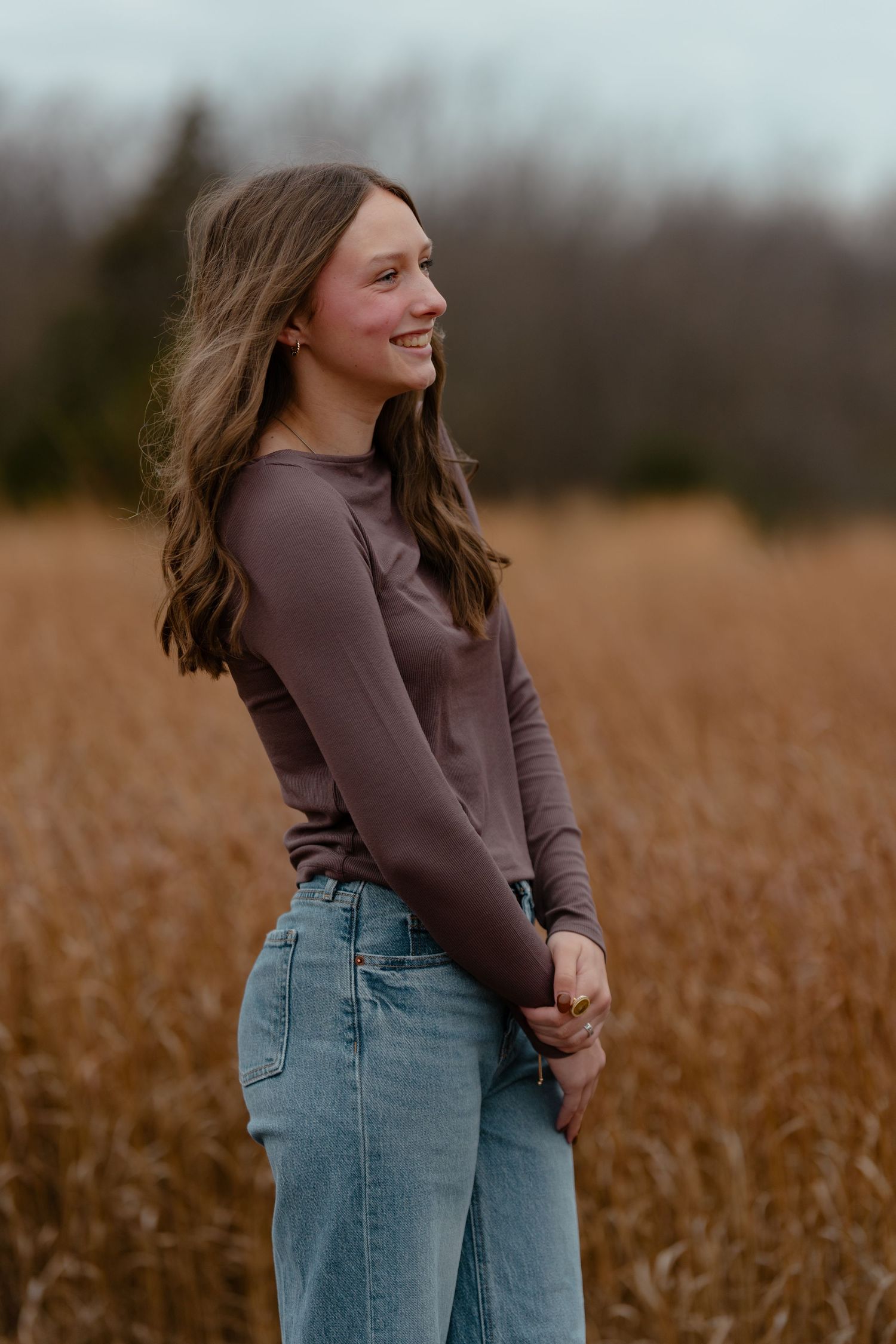 Young woman in brown long-sleeve top and jeans smiling in a golden field with trees in background.