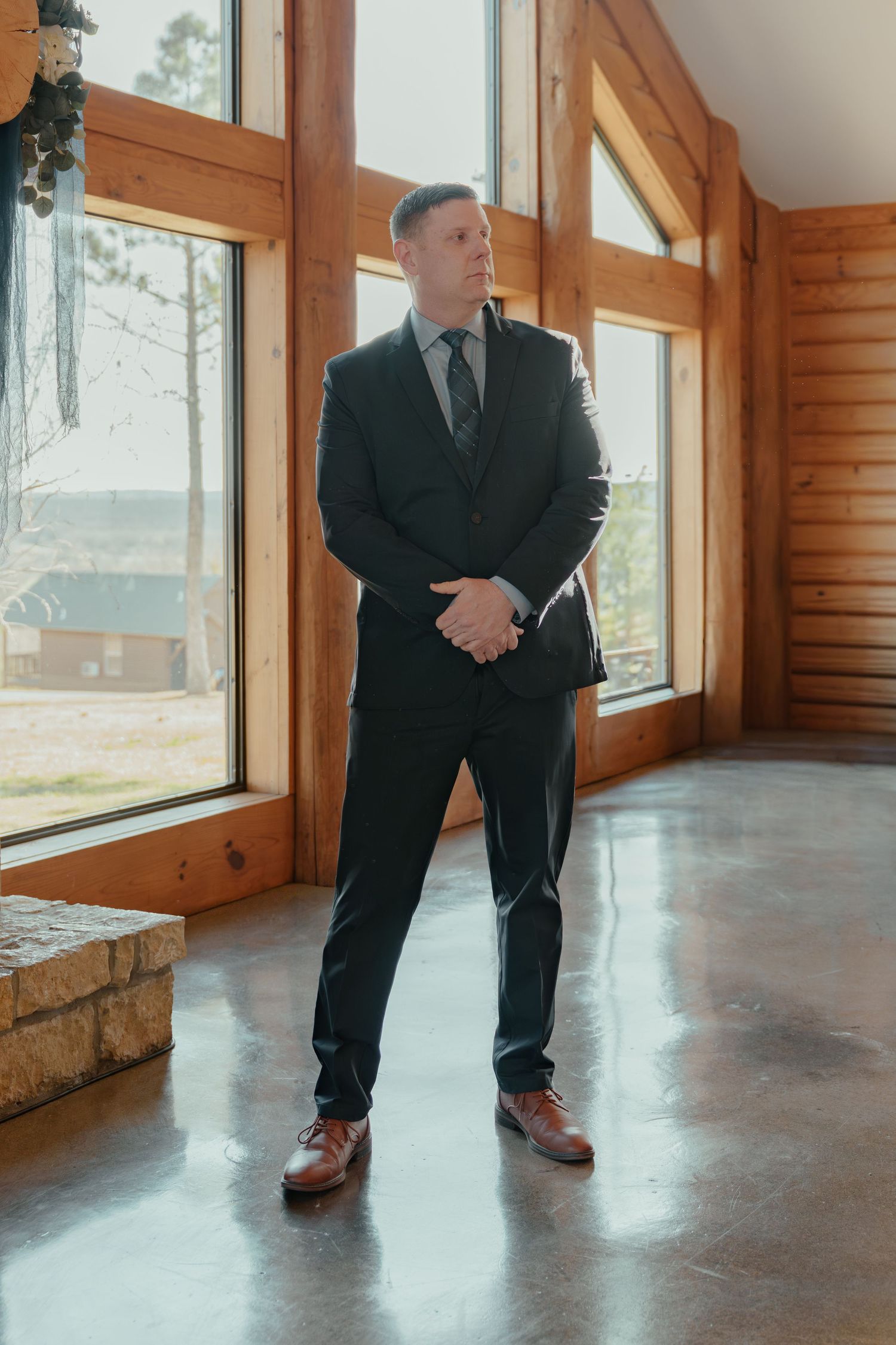 Man in dark suit and brown dress shoes standing in a rustic log cabin venue with large windows.