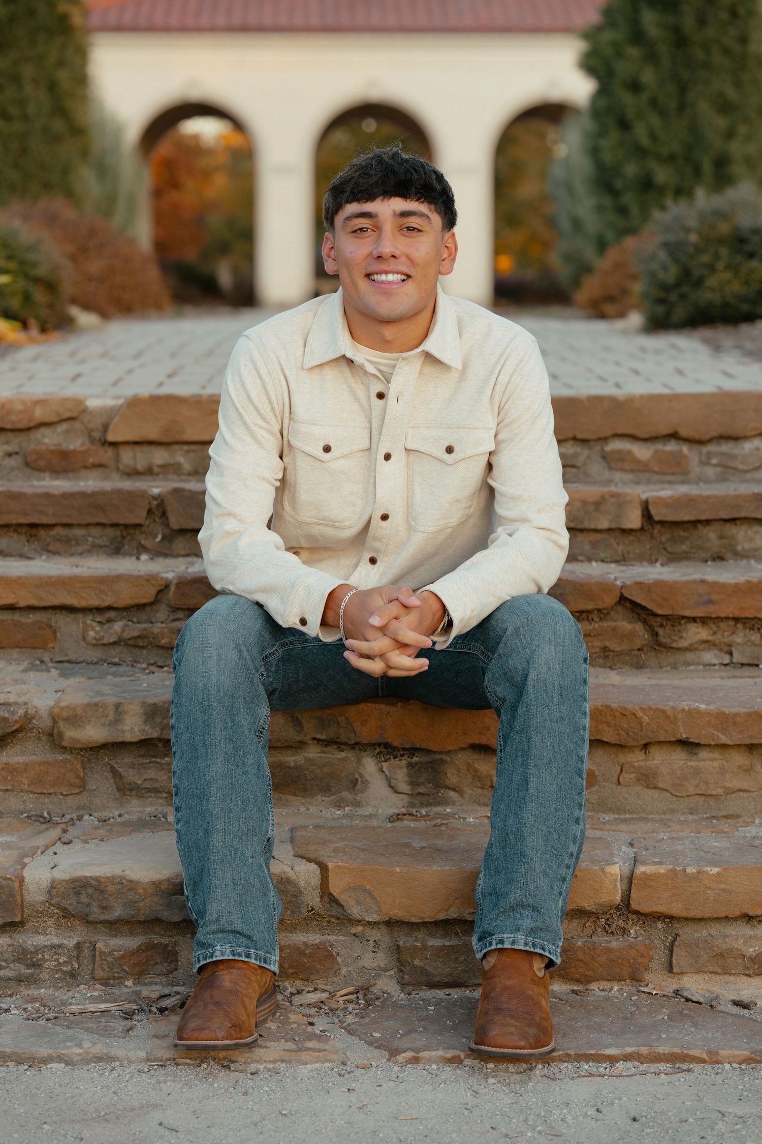 A person in a cream jacket and jeans sits casually on stone steps with arched architecture in the background.