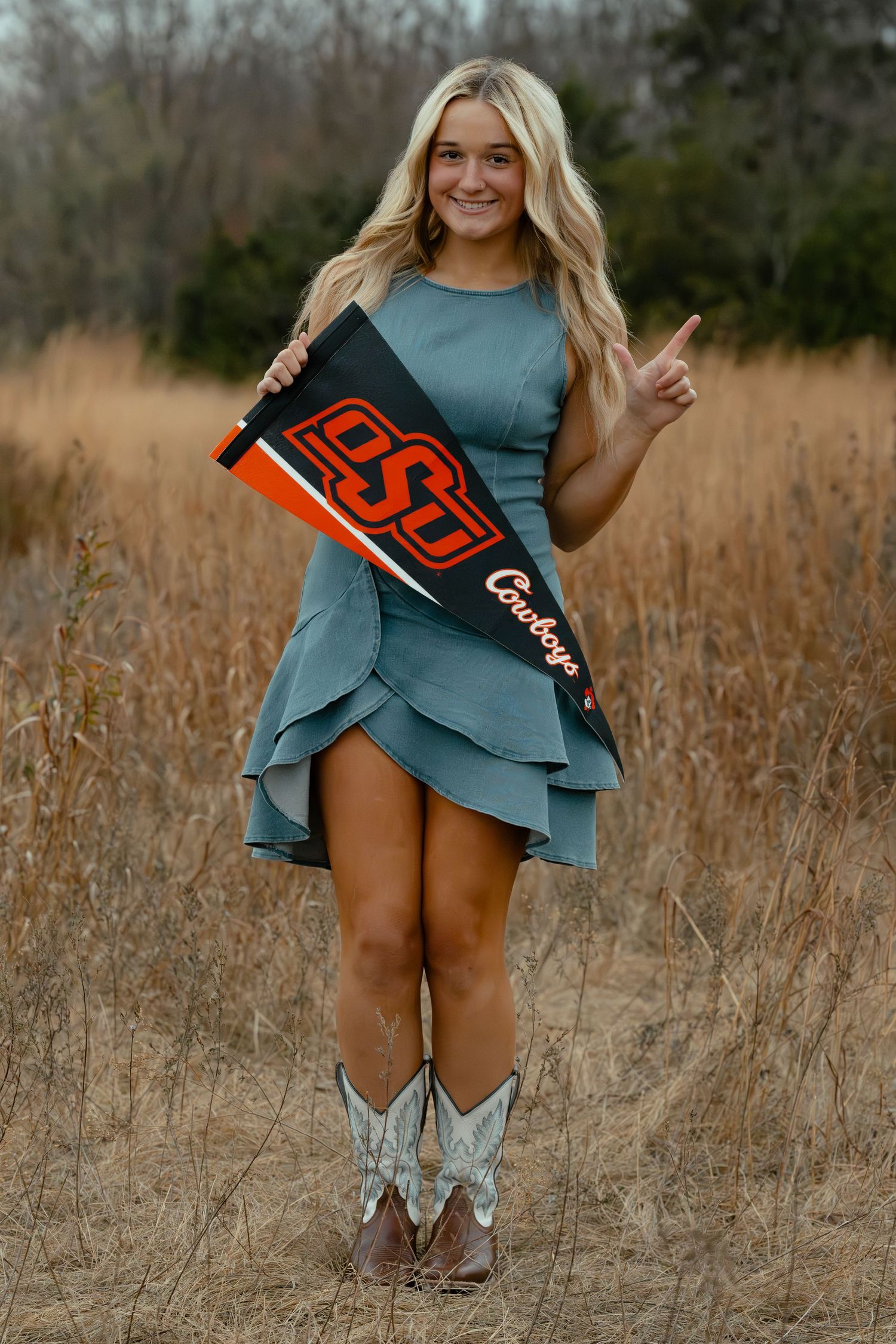 Blonde woman in teal dress and cowboy boots holding OSU Cowboys pennant in a field.