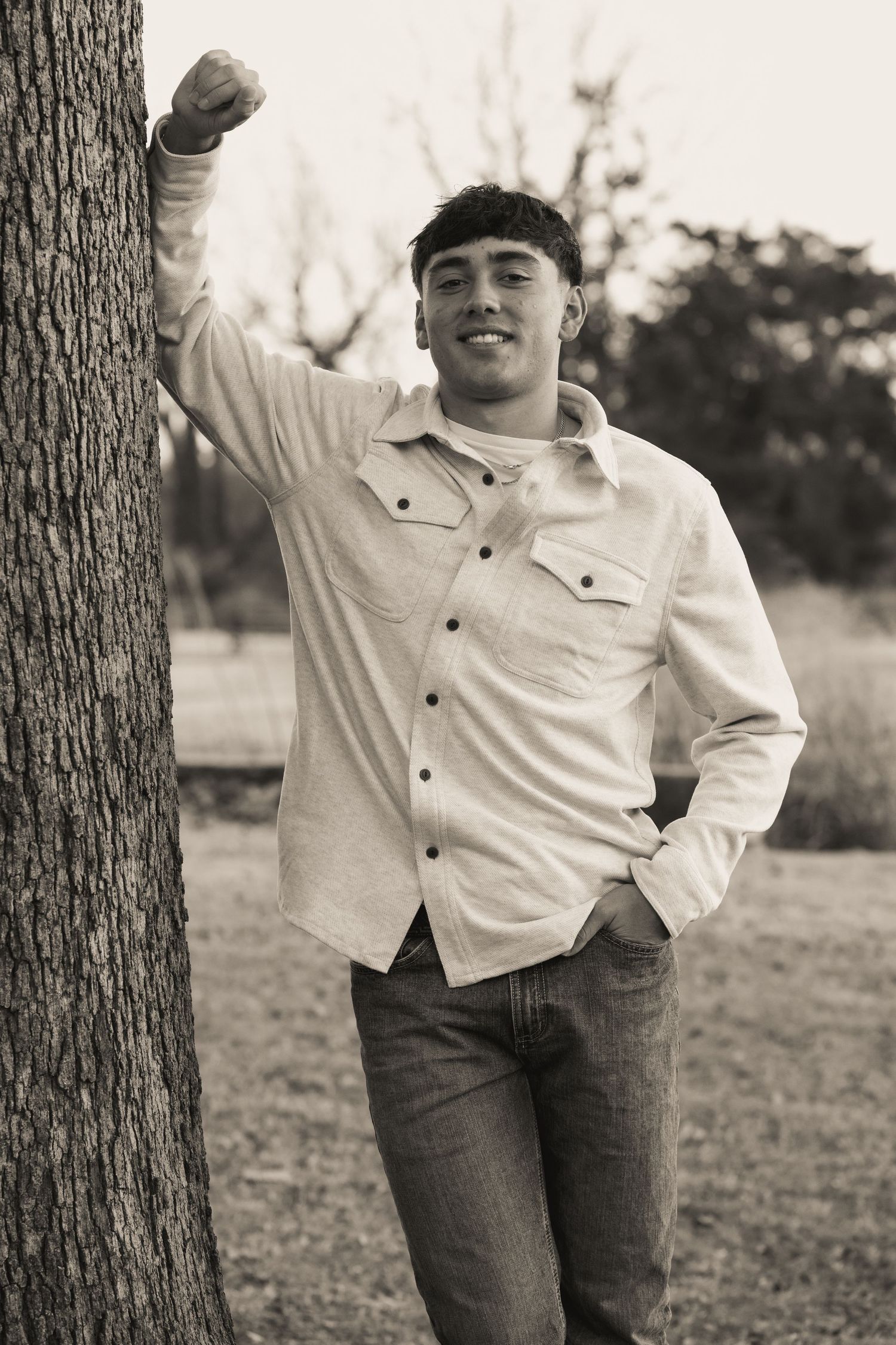 Black and white portrait of someone in casual attire leaning against tree outdoors.