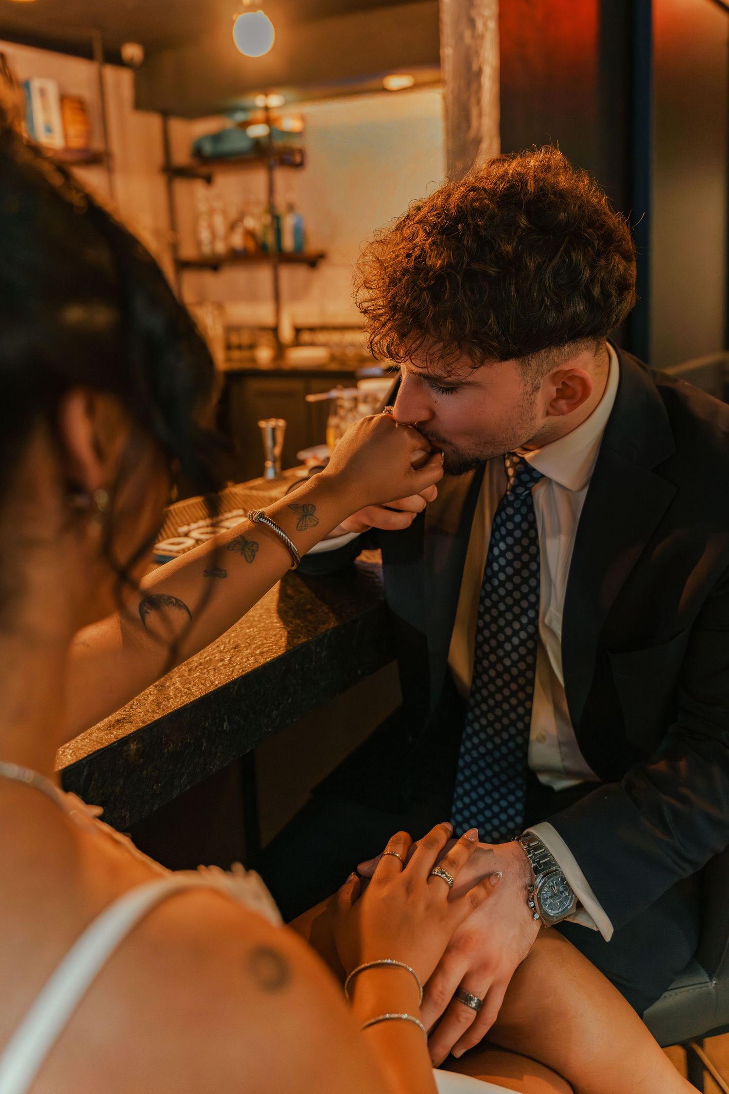 Groom in navy suit kisses bride's hand tenderly at a warmly lit restaurant during their wedding reception.