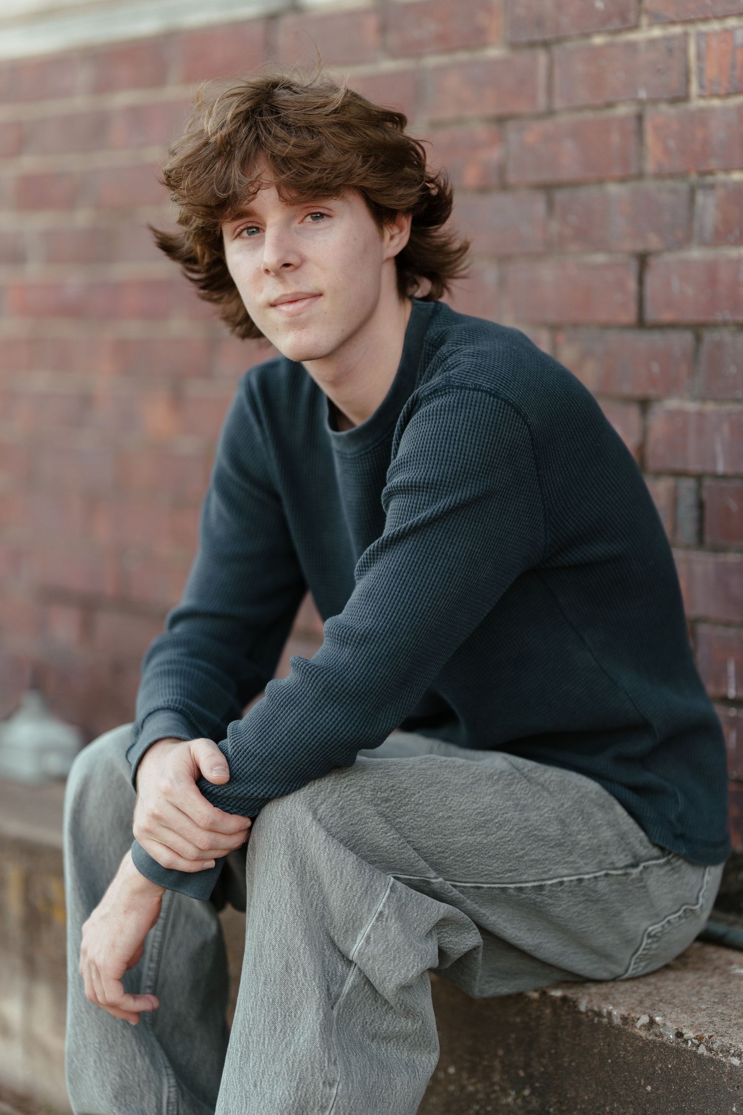 Young man with curly hair sitting on a ledge in a dark sweater and gray jeans against a brick wall.