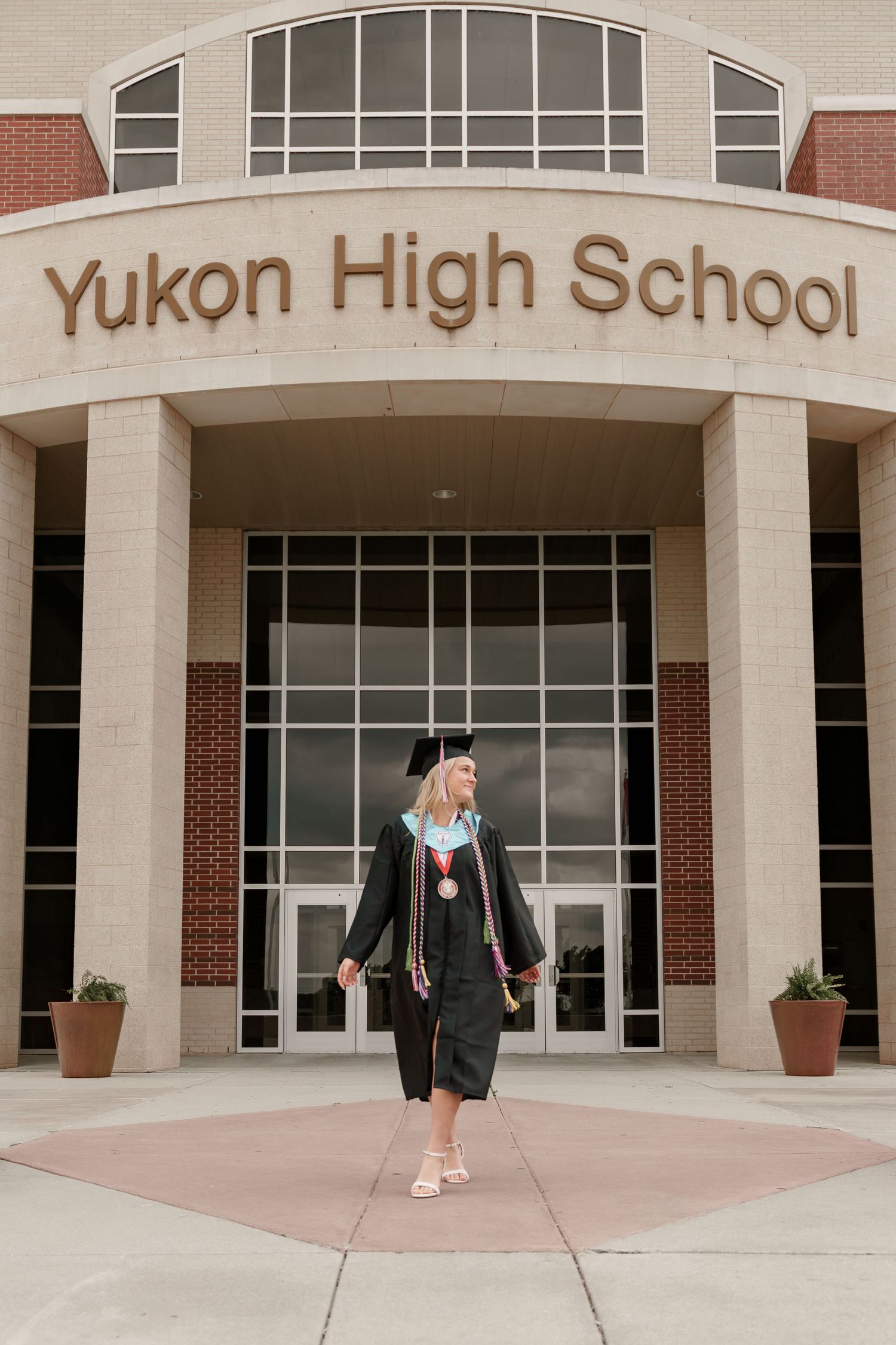 A graduating student in cap and gown walks proudly in front of Yukon High School entrance on graduation day.