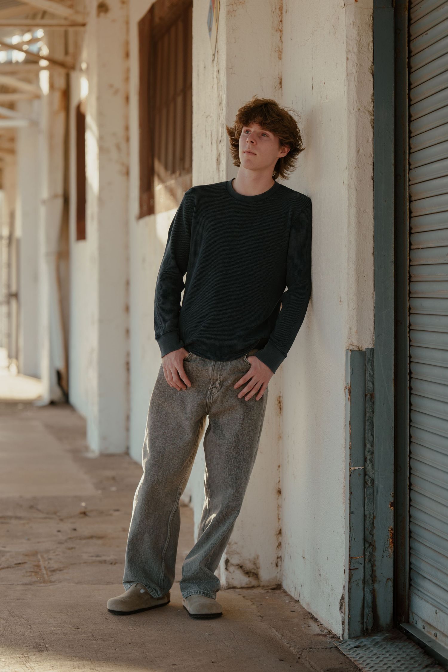 Young man in dark green sweatshirt and gray jeans leaning against weathered white wall in urban corridor.
