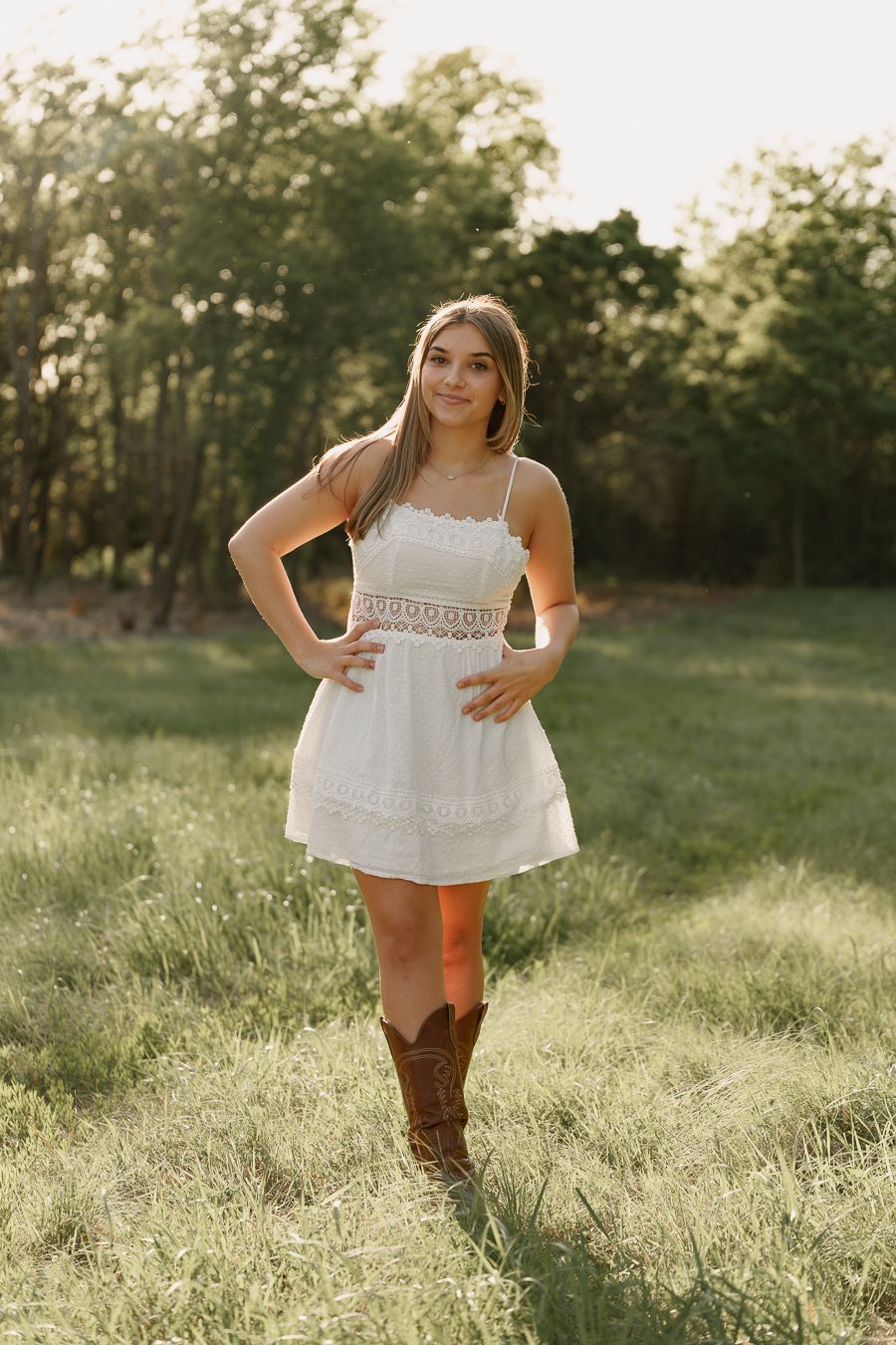 Young woman in white lace dress and brown cowboy boots posing in a sunlit green field.