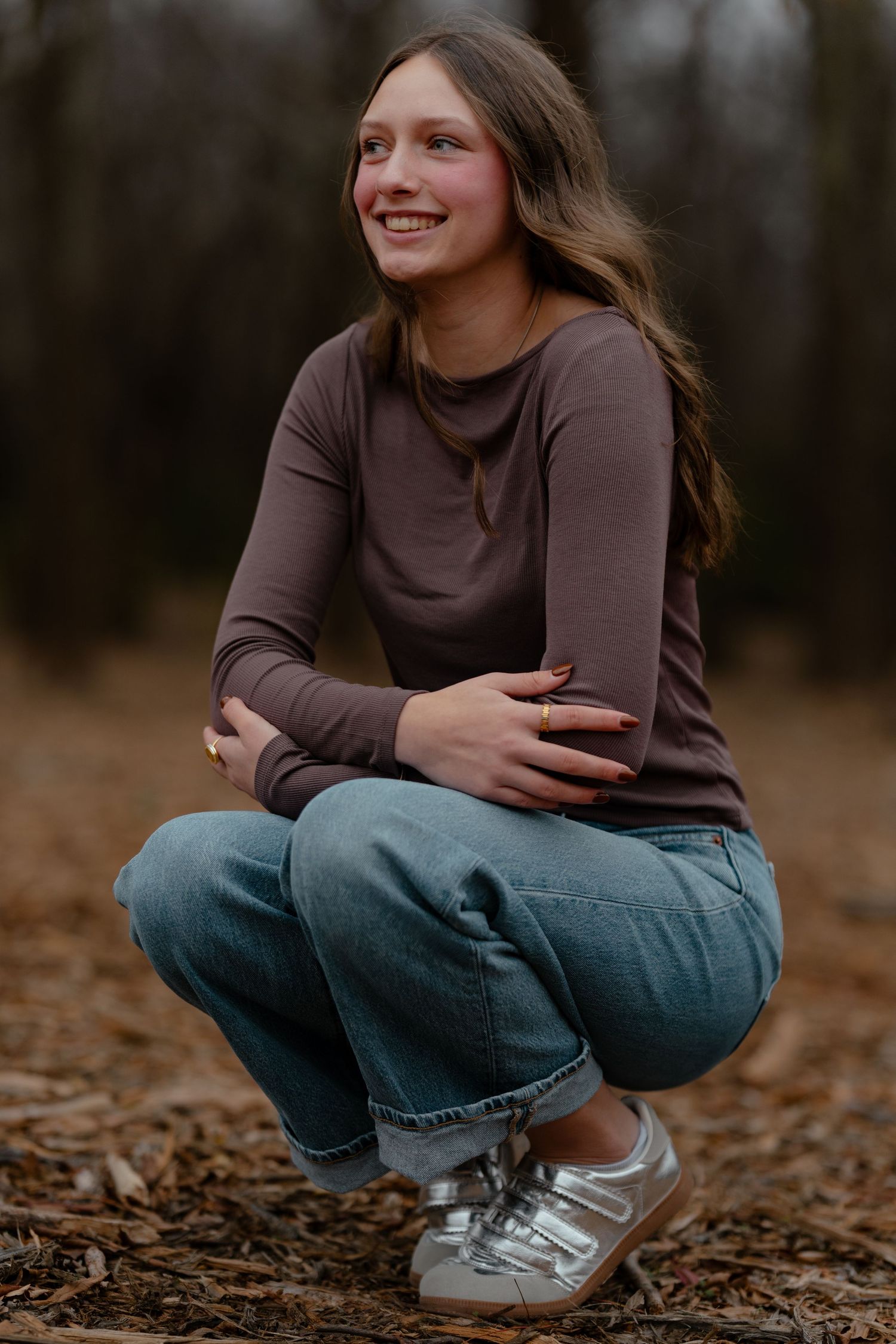 Smiling teenage girl crouching outdoors in brown top, blue jeans, and silver sneakers on a leaf-covered forest path.