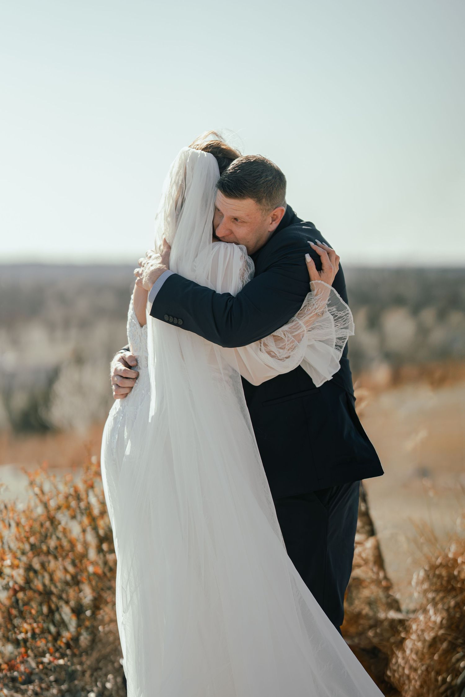 Bride and groom embracing outdoors on their wedding day, with autumn landscape in background.