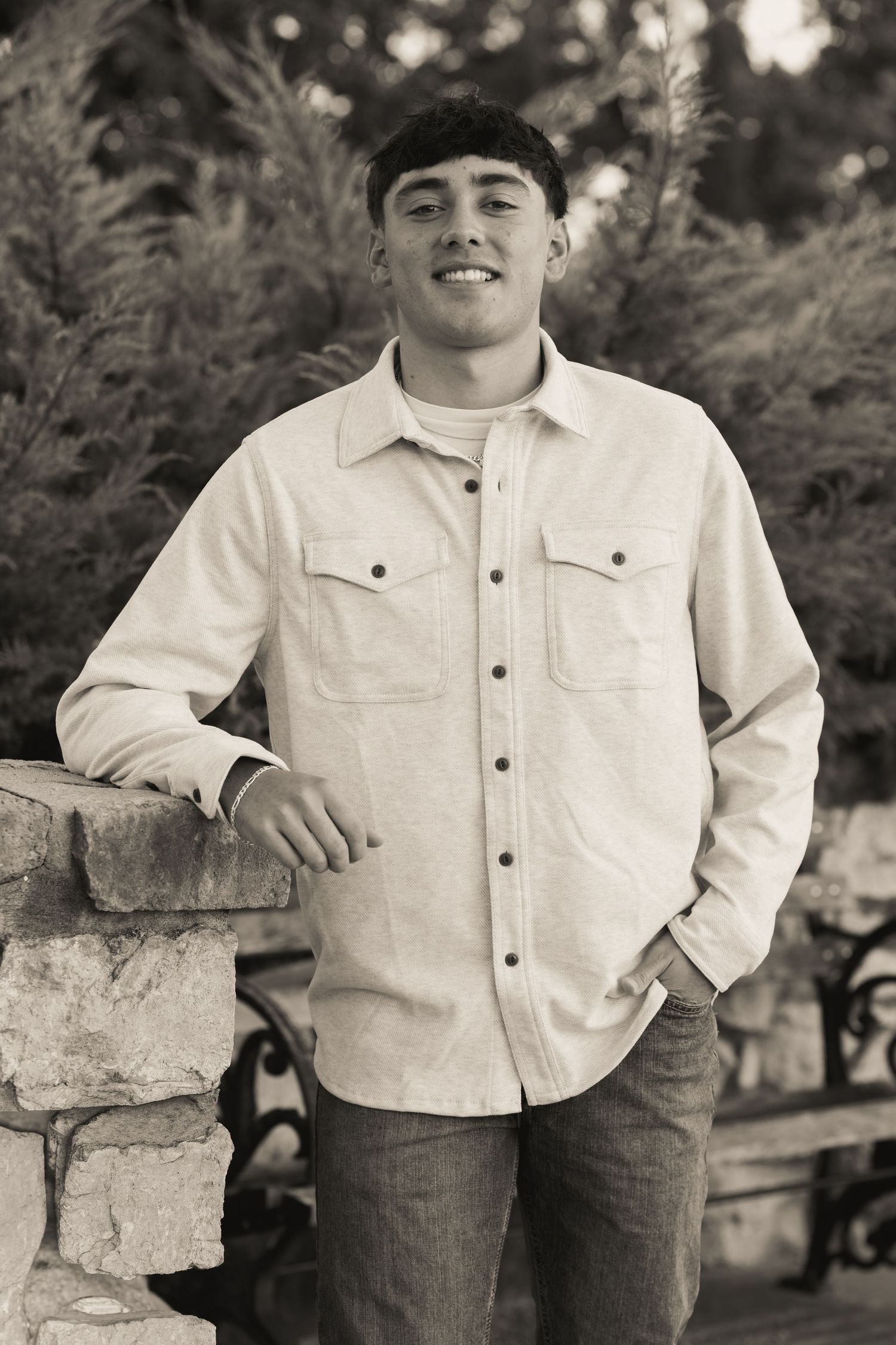 A black and white portrait of someone in a light jacket leaning against a stone wall outdoors.
