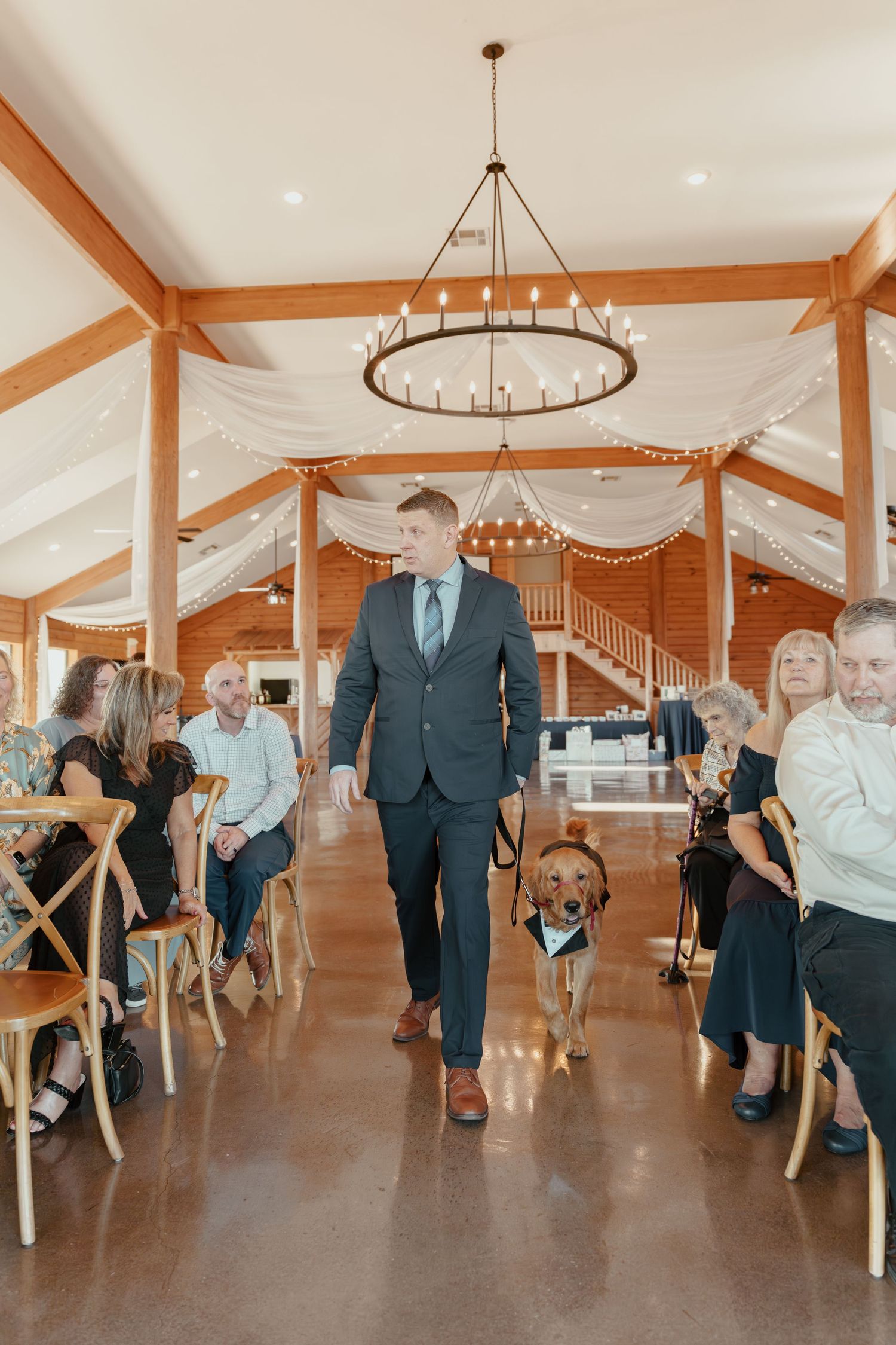 Groom walks dog down wedding aisle in elegant barn venue with wooden beams and draped white fabric.