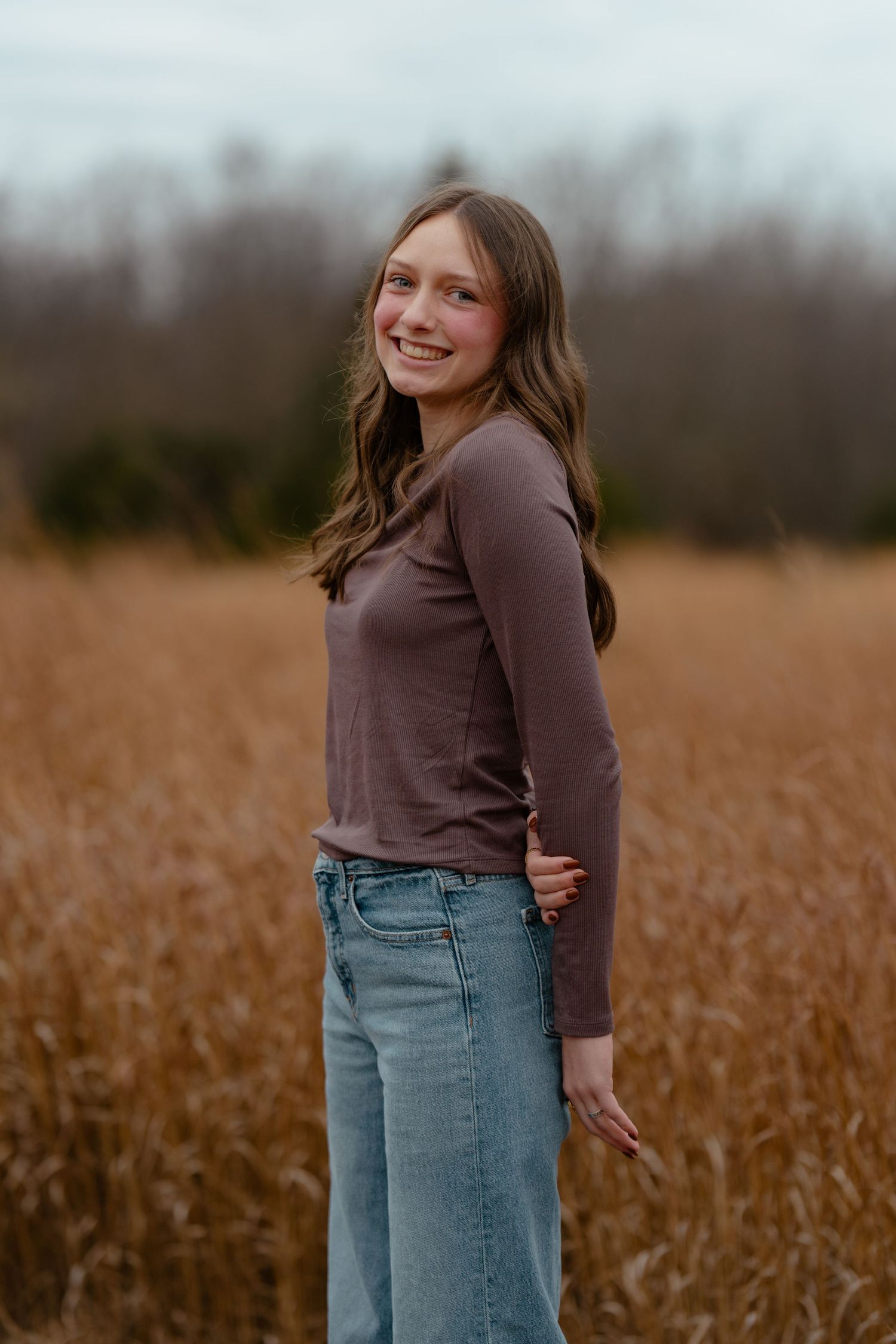 Smiling teenage girl in brown long-sleeve top and wide-leg jeans posing in a golden field.
