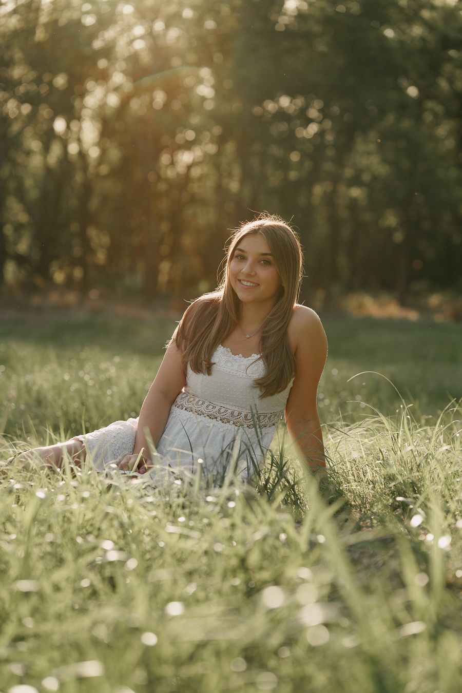 Young woman in white dress sitting in sunlit green meadow with trees in background, golden hour portrait photography.