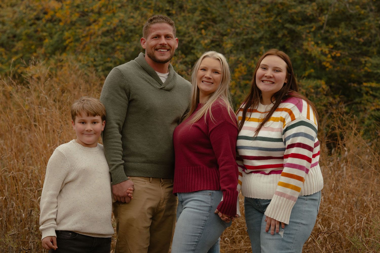 Happy family of four wearing warm sweaters poses together in autumn field.