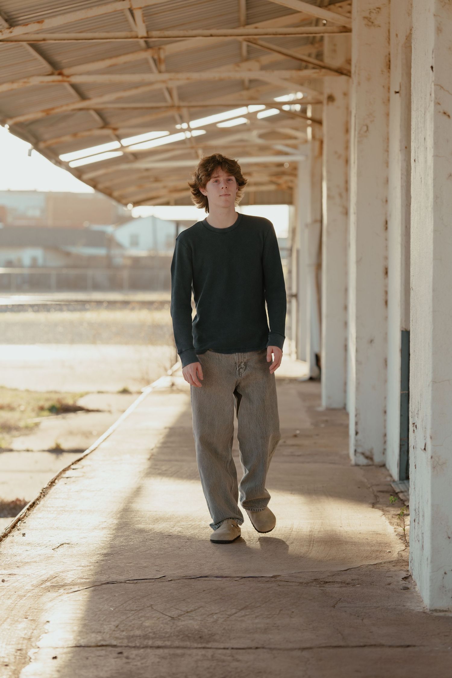 Young man with curly hair walking under a covered outdoor walkway, wearing a dark green sweater and gray sweatpants.