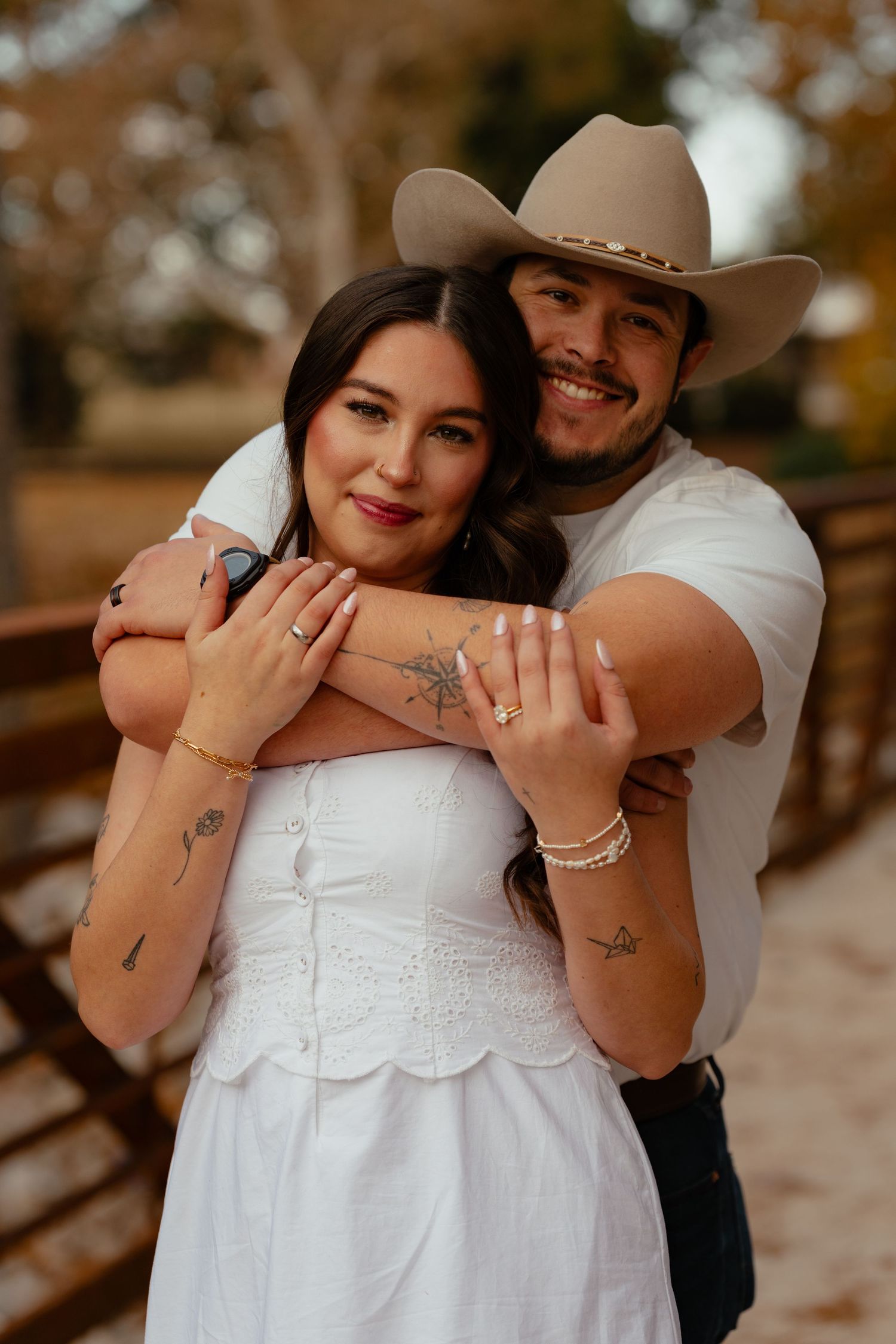 Romantic couple embraces outdoors with woman in white dress and man in cowboy hat during autumn photoshoot.