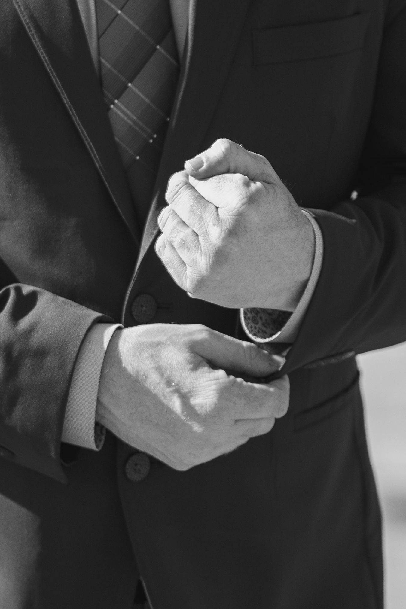Black and white photo of a man in a suit adjusting his cufflinks, wearing a watch and striped tie.