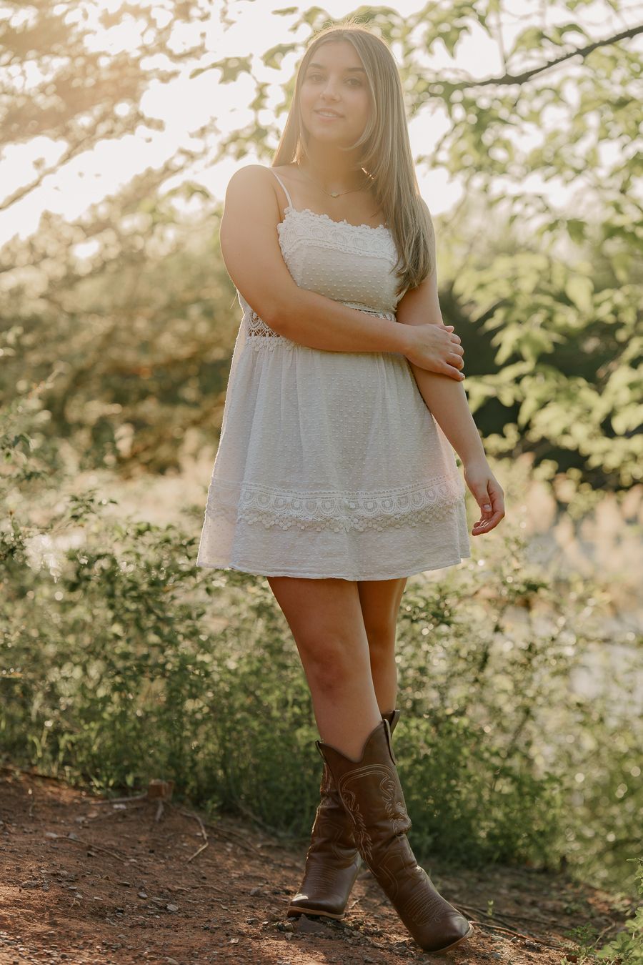 Young woman in white lace mini dress and brown cowboy boots standing outdoors in golden sunlight.