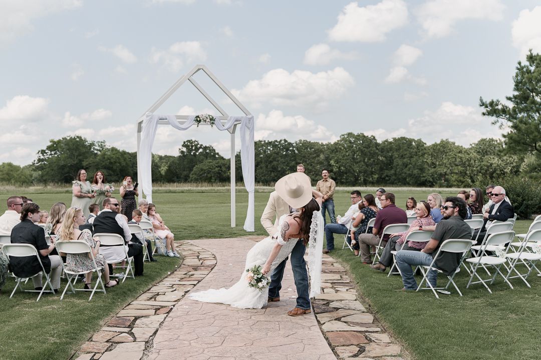 Couple shares first kiss at outdoor wedding ceremony with white arch, stone path aisle, and seated guests on a sunny day.