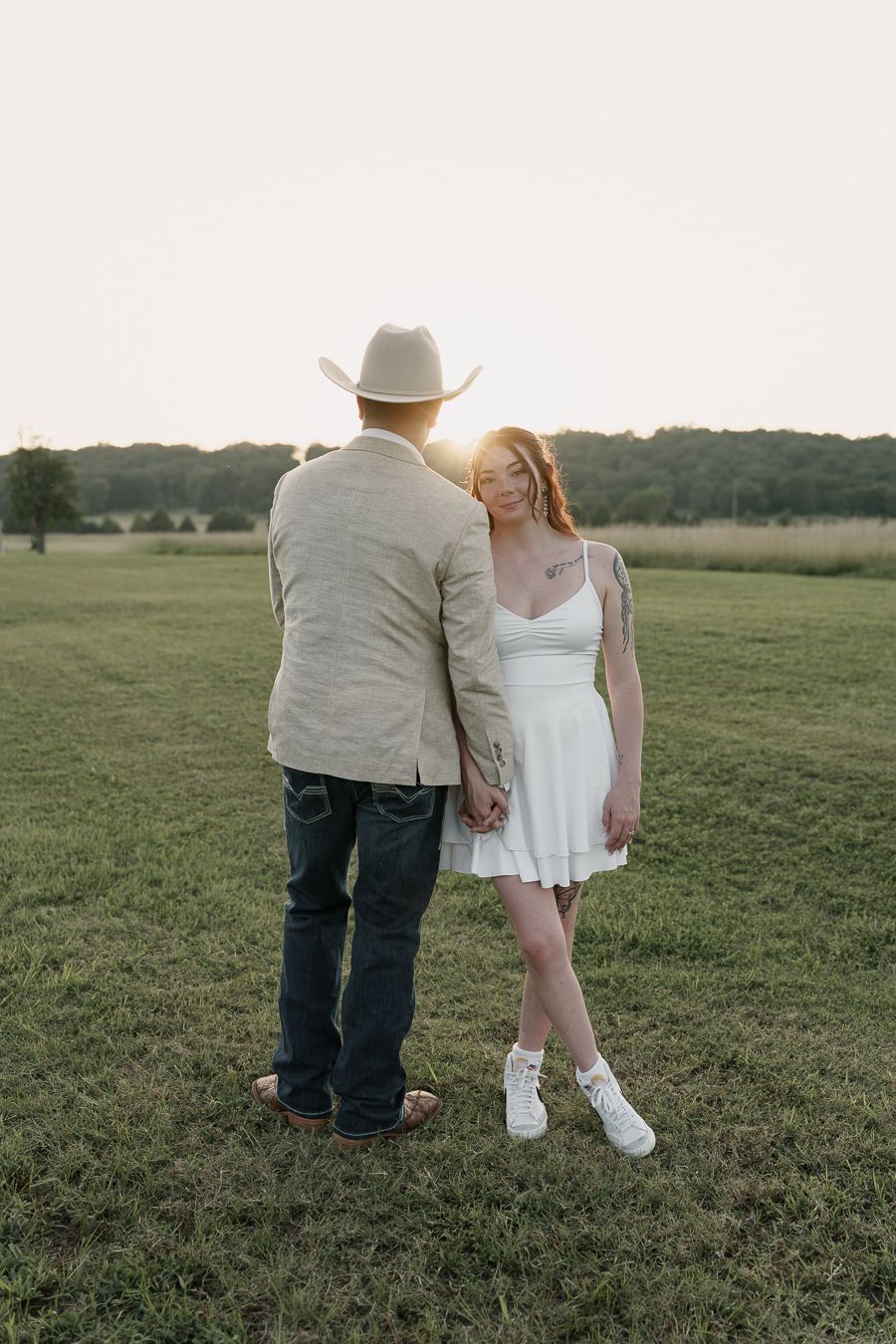 Couple holding hands at golden hour in a green field, woman in white dress and man in cowboy hat and jeans.