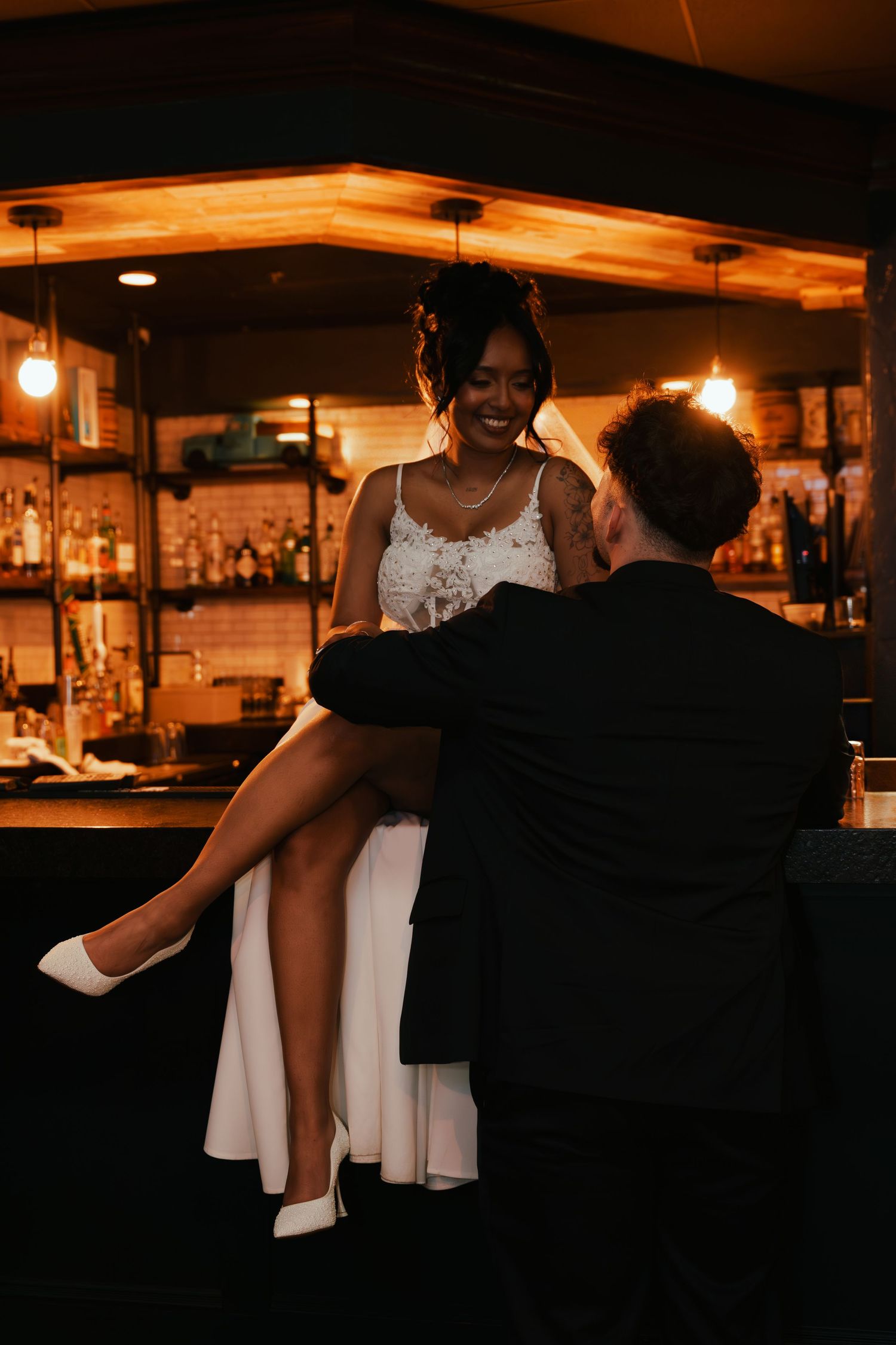 A couple shares an intimate moment at a dimly lit bar, with warm lighting and bottles displayed in the background.