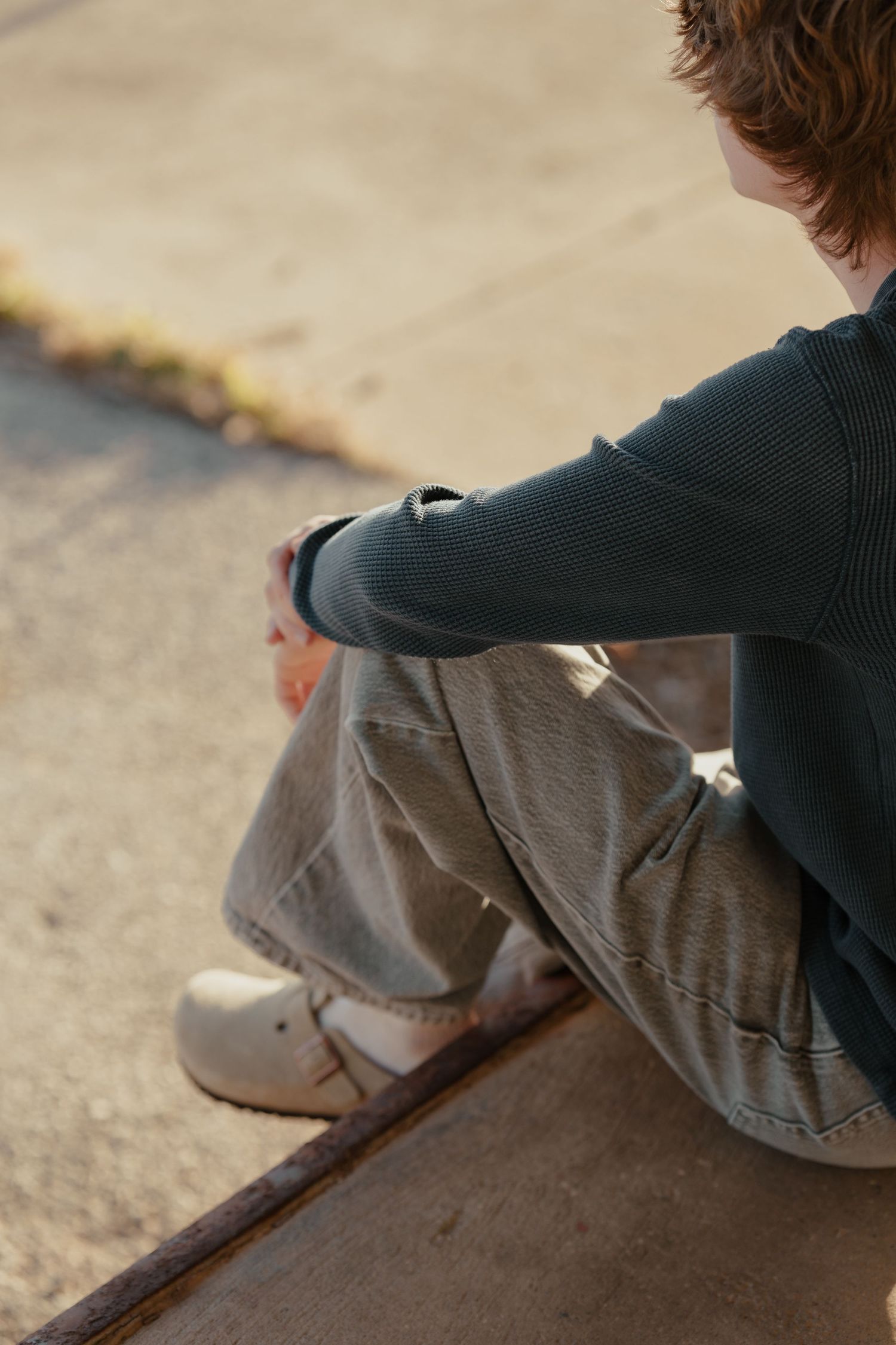 A young person sits on a curb outdoors, wearing khaki pants, a dark top, and white shoes, viewed from behind.