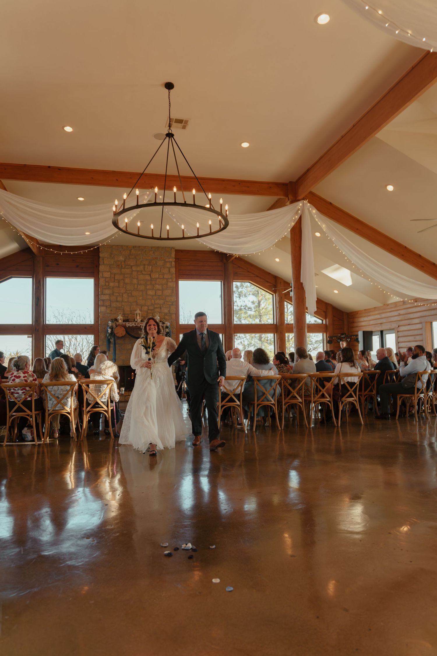 Spacious wedding reception hall with stone walls, wooden beams, and circular chandeliers features crossback chairs.