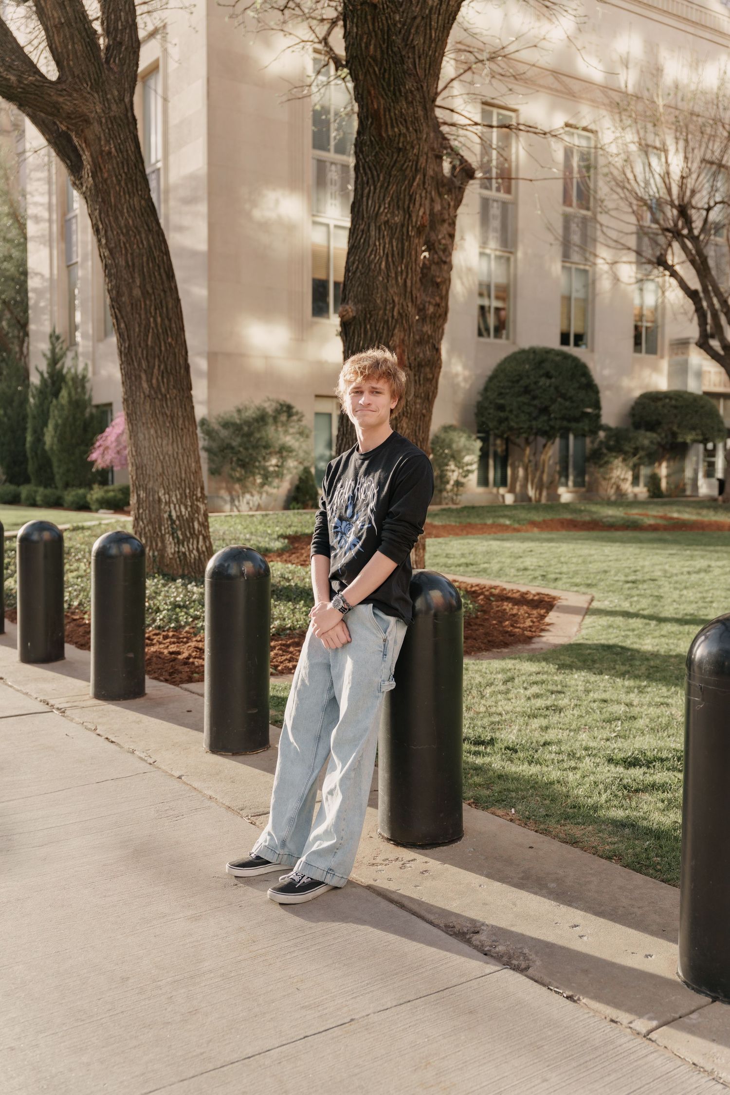 Young man in black graphic tee and wide-leg jeans leaning on bollard outside a classic brick building.