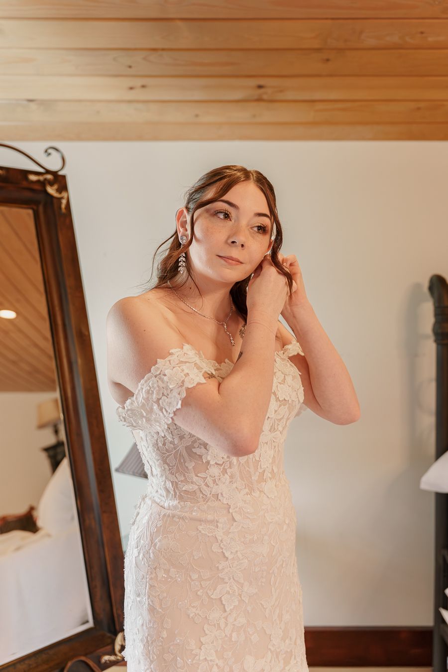 Bride in off-shoulder lace wedding gown adjusting earrings near ornate mirror in warmly lit bridal suite.