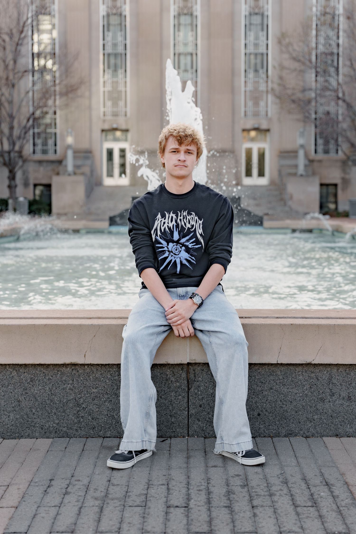 Young man in black graphic sweatshirt and jeans sitting on fountain ledge in front of a large building.