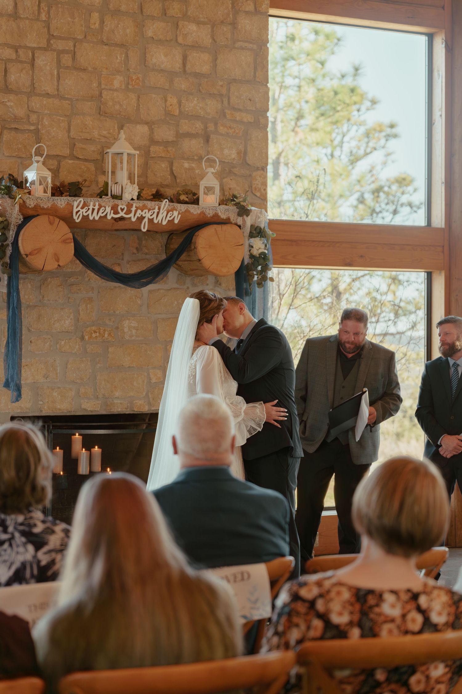 Bride and groom share first kiss at rustic stone fireplace wedding ceremony with Better Together sign.