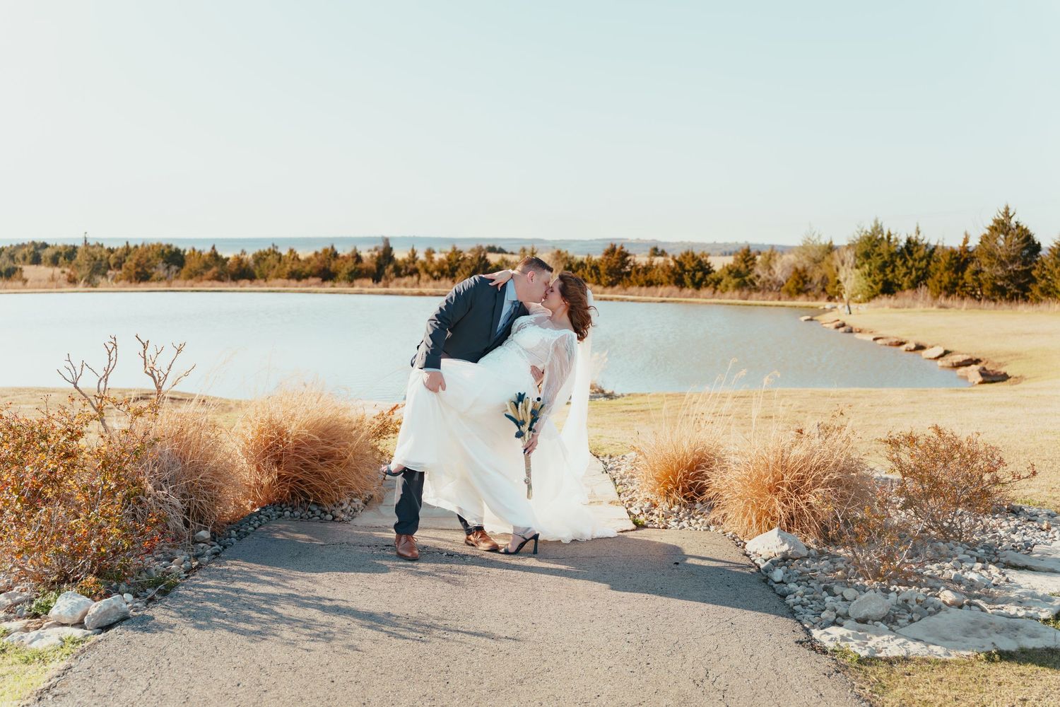 Bride and groom share a romantic dip kiss on a scenic lakeside path surrounded by golden autumn shrubs.