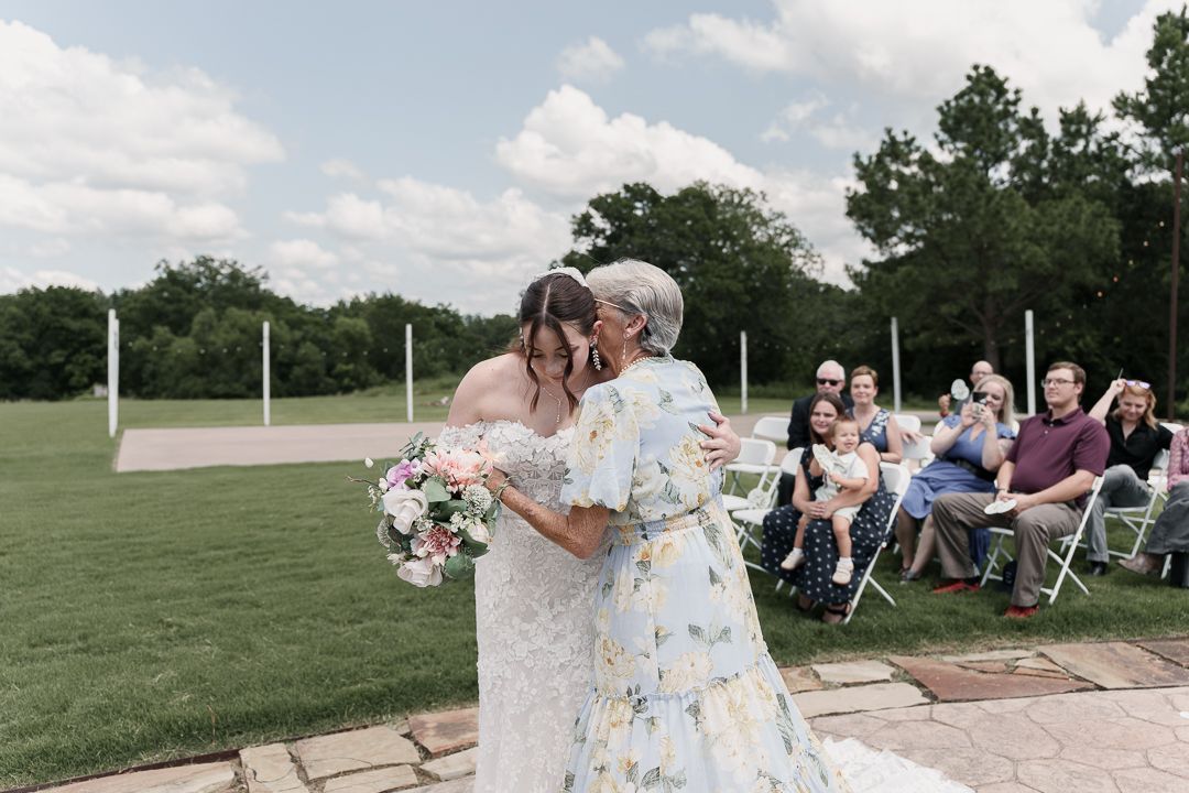 Two brides share a kiss during outdoor wedding ceremony on green lawn with seated guests.