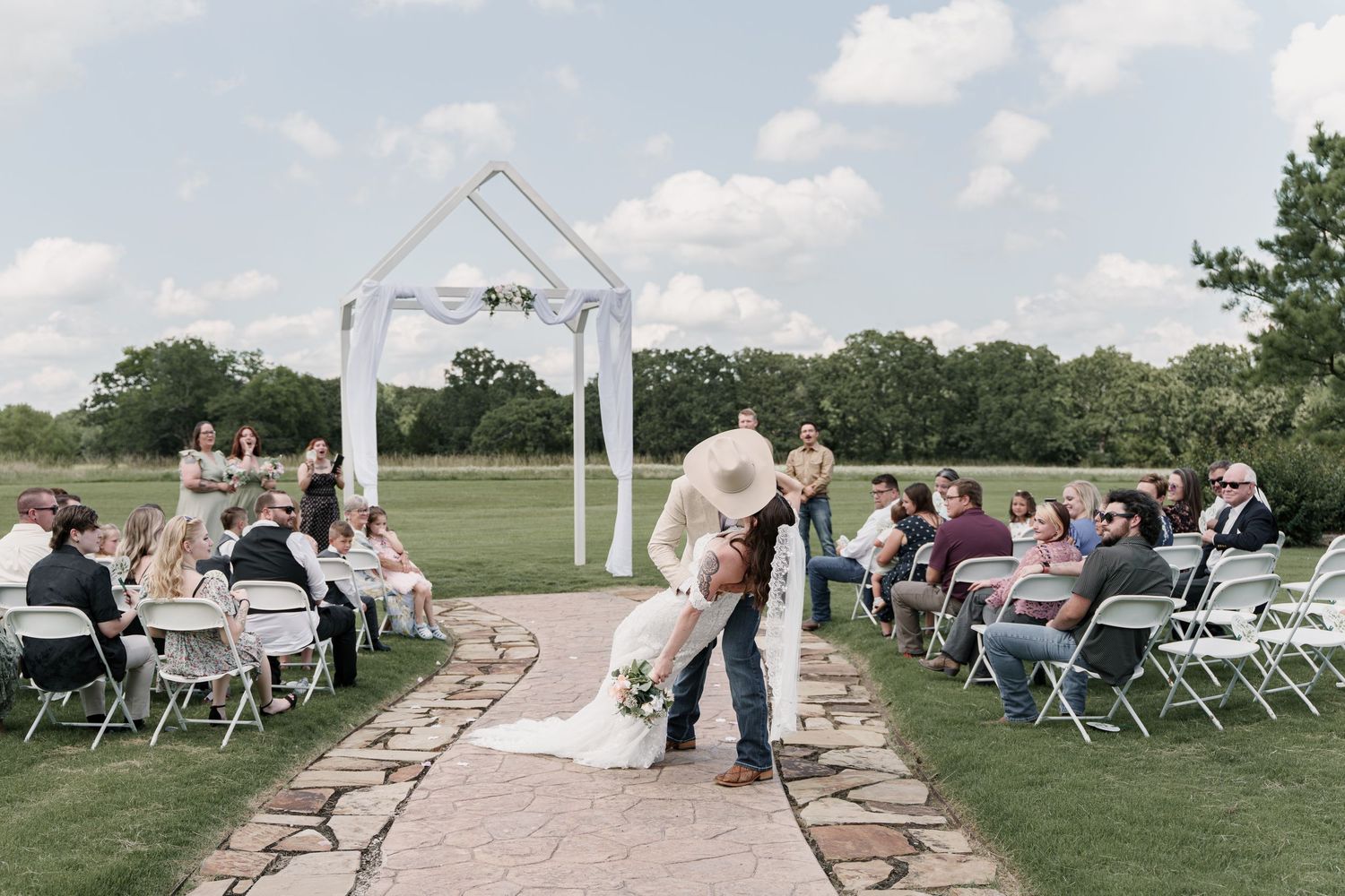 Outdoor wedding ceremony on stone path with couple kissing under white geometric arch, guests seated on white chairs on green lawn.