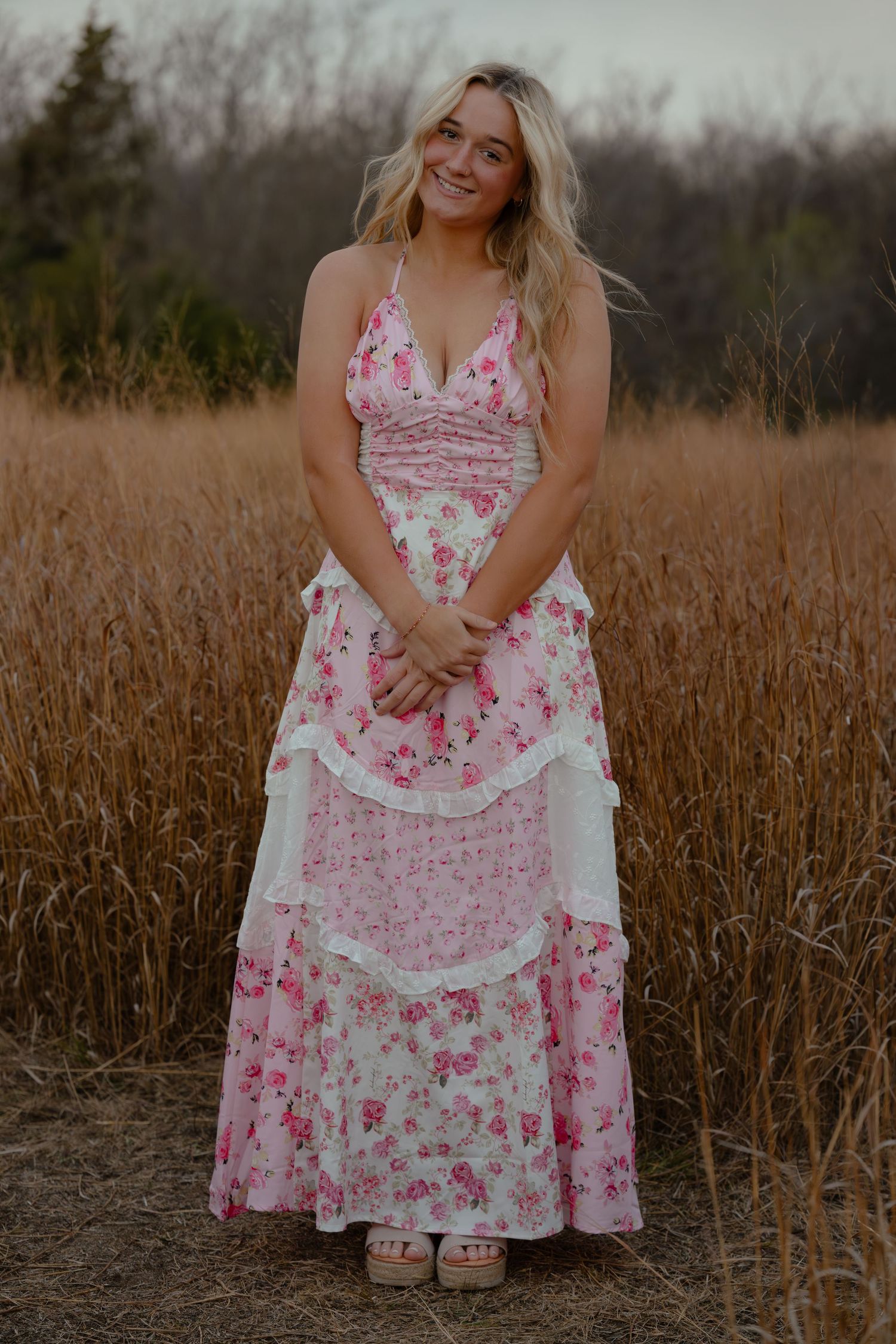 Blonde woman in pink floral tiered maxi dress standing in golden wheat field, smiling outdoors.