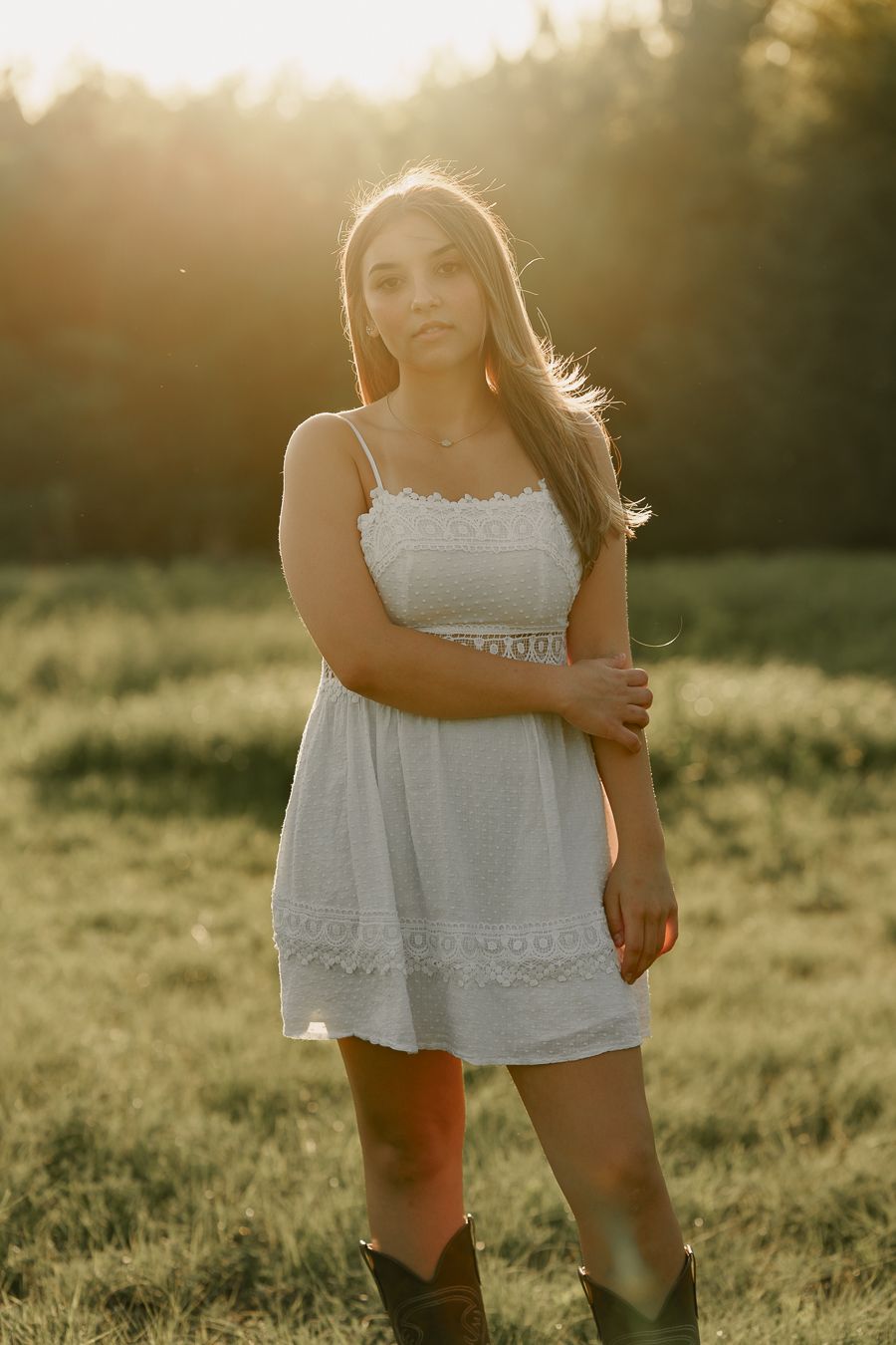 Young woman in white sundress and cowboy boots posing in golden sunset field.