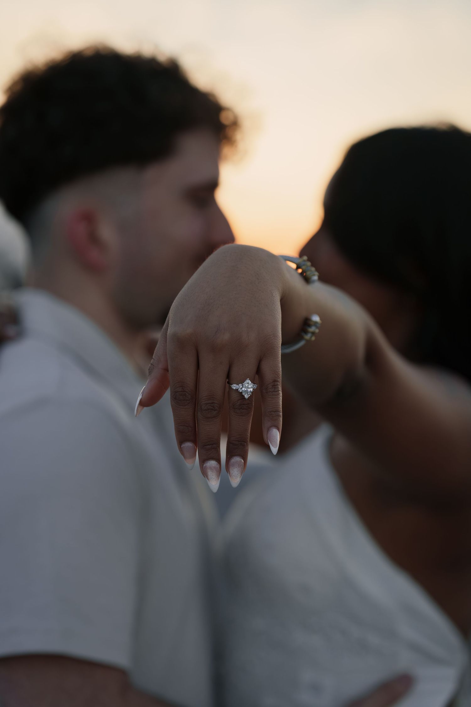 Close-up view of an engagement ring on someone's hand during a sunset gathering with people in white attire.