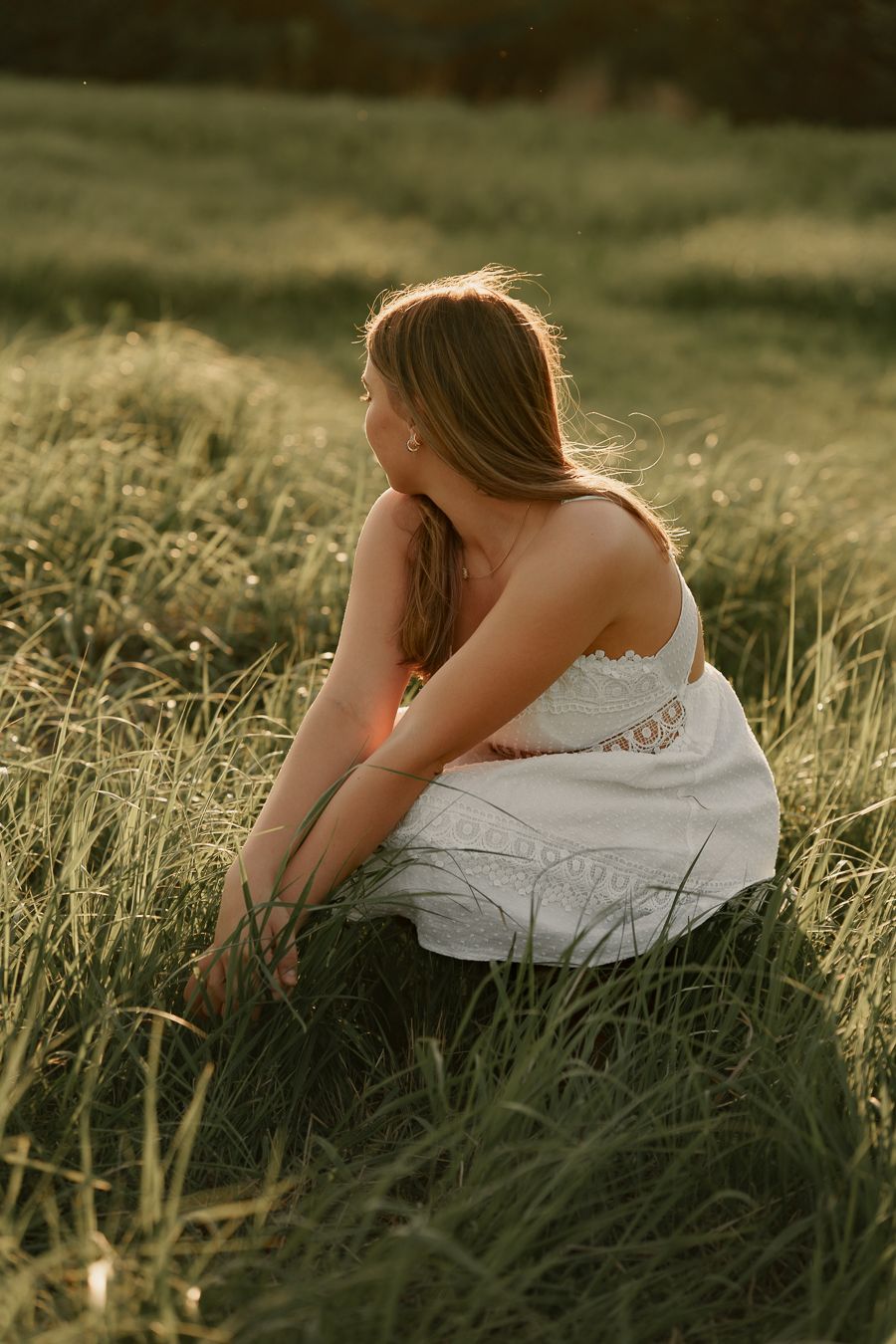 Young woman in white sundress sitting in tall green grass during golden hour sunset.