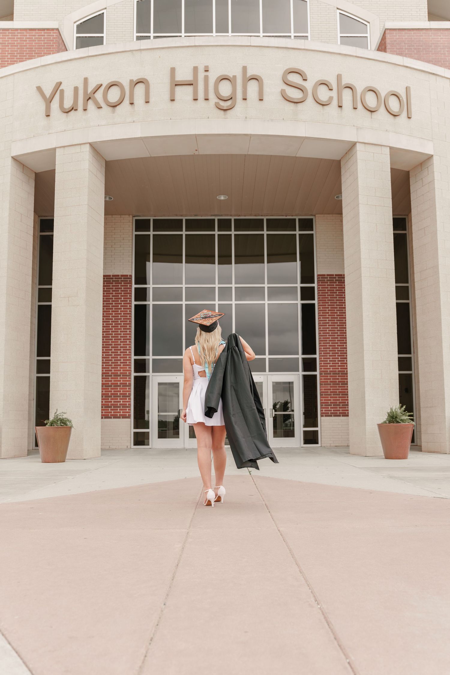 A graduating student walks toward Yukon High School entrance wearing cap and gown in white dress and heels.
