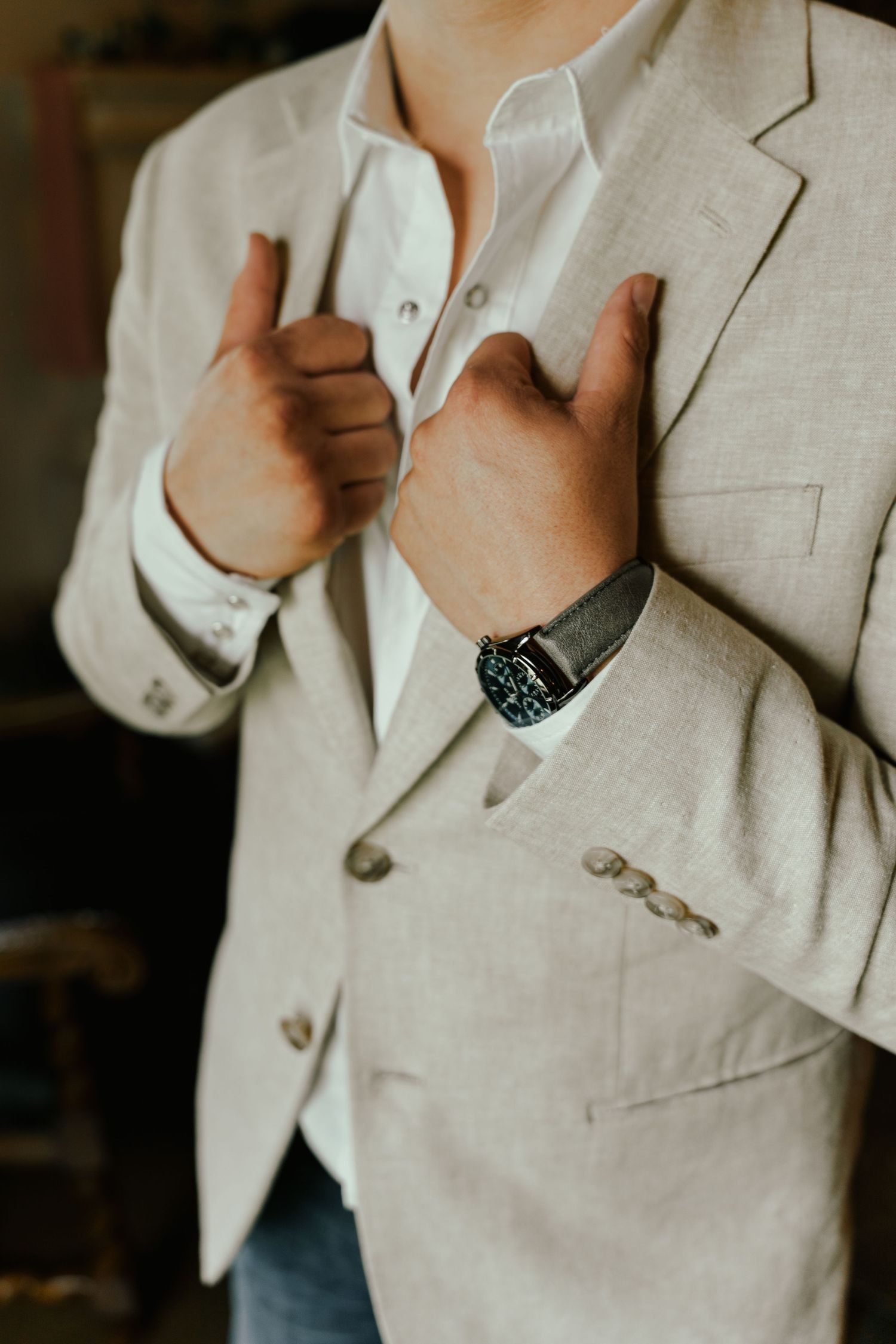 Man in beige linen blazer adjusting lapels, wearing a stylish wristwatch, semi-formal wedding attire.