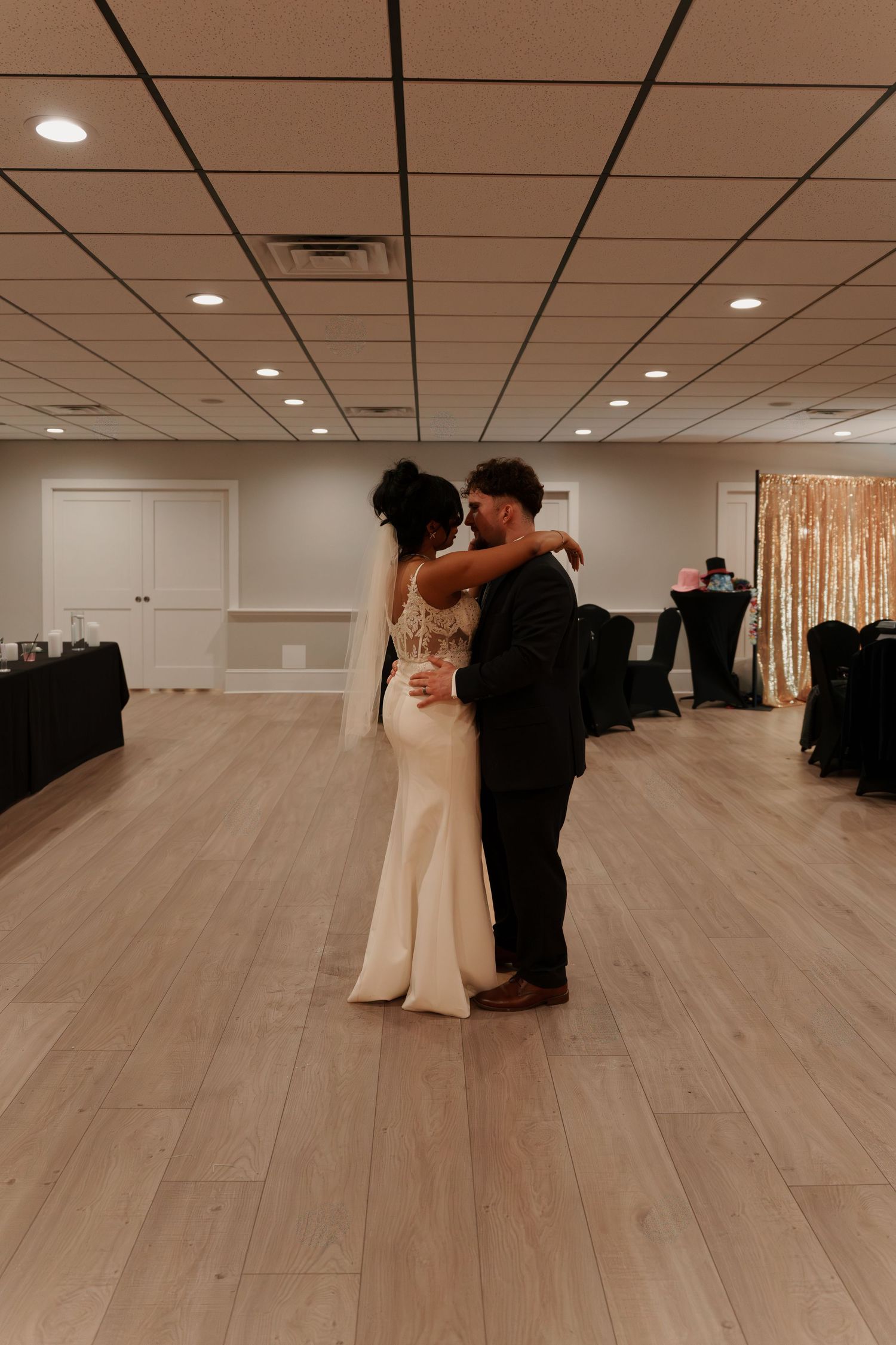 A couple dances together in an elegant ballroom with light wooden floors and recessed lighting overhead.