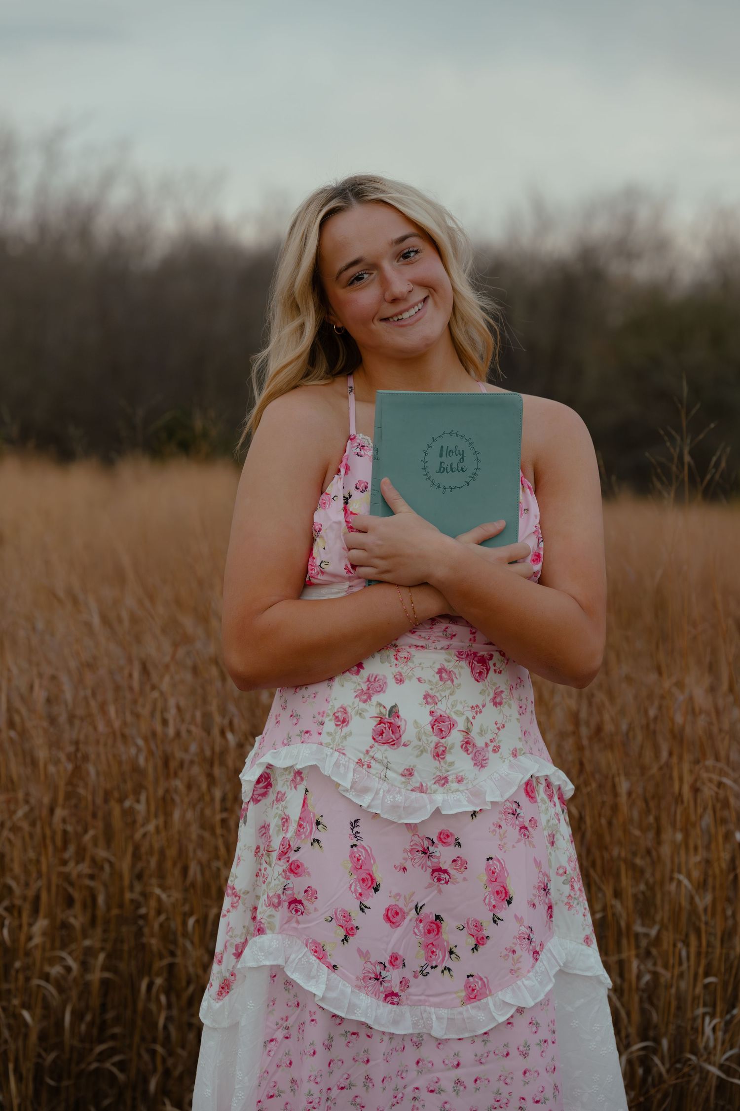 Person in floral pink dress holds teal book while standing in tall grass field.