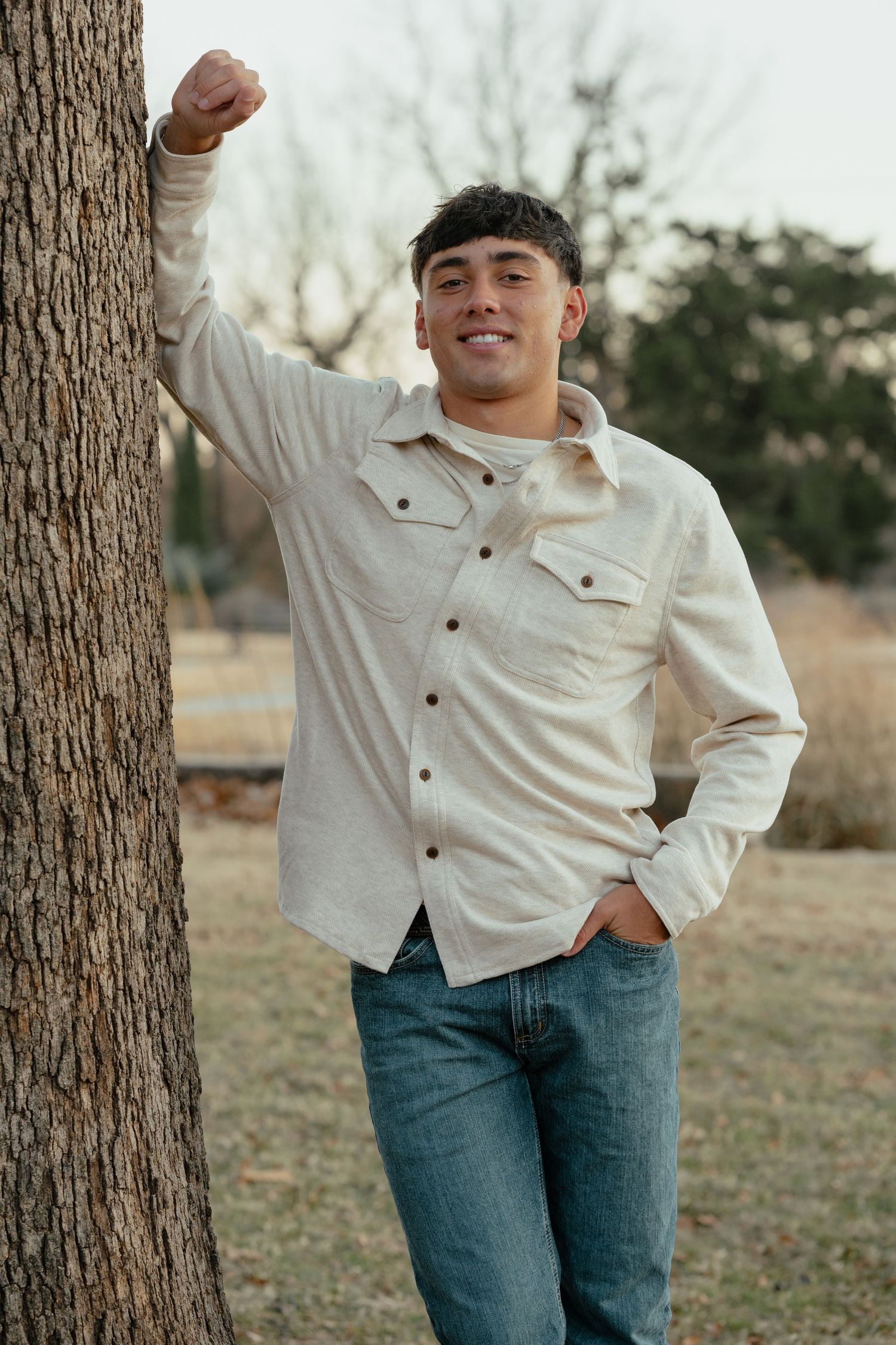 Casual outdoor portrait of someone leaning against a tree wearing a cream button-down shirt and blue jeans.