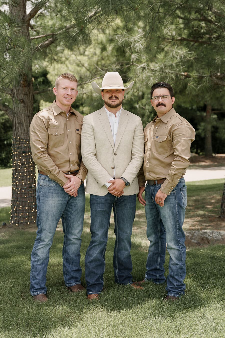 Three men in Western attire with jeans and tan shirts pose outdoors, center man wearing a cowboy hat and blazer.