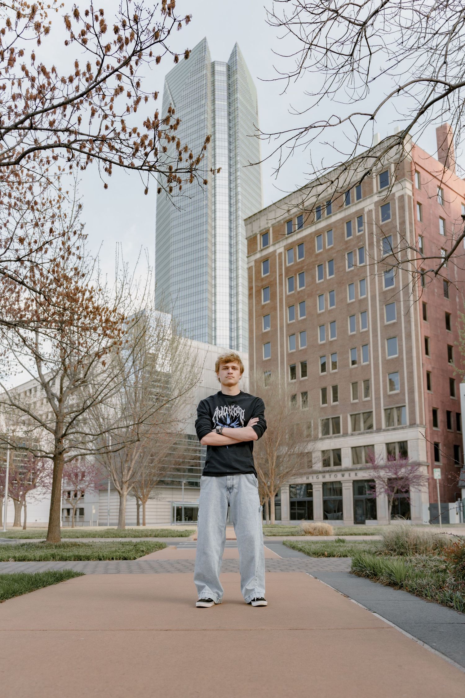 Young man standing confidently with arms crossed on urban pathway surrounded by city buildings and bare trees.