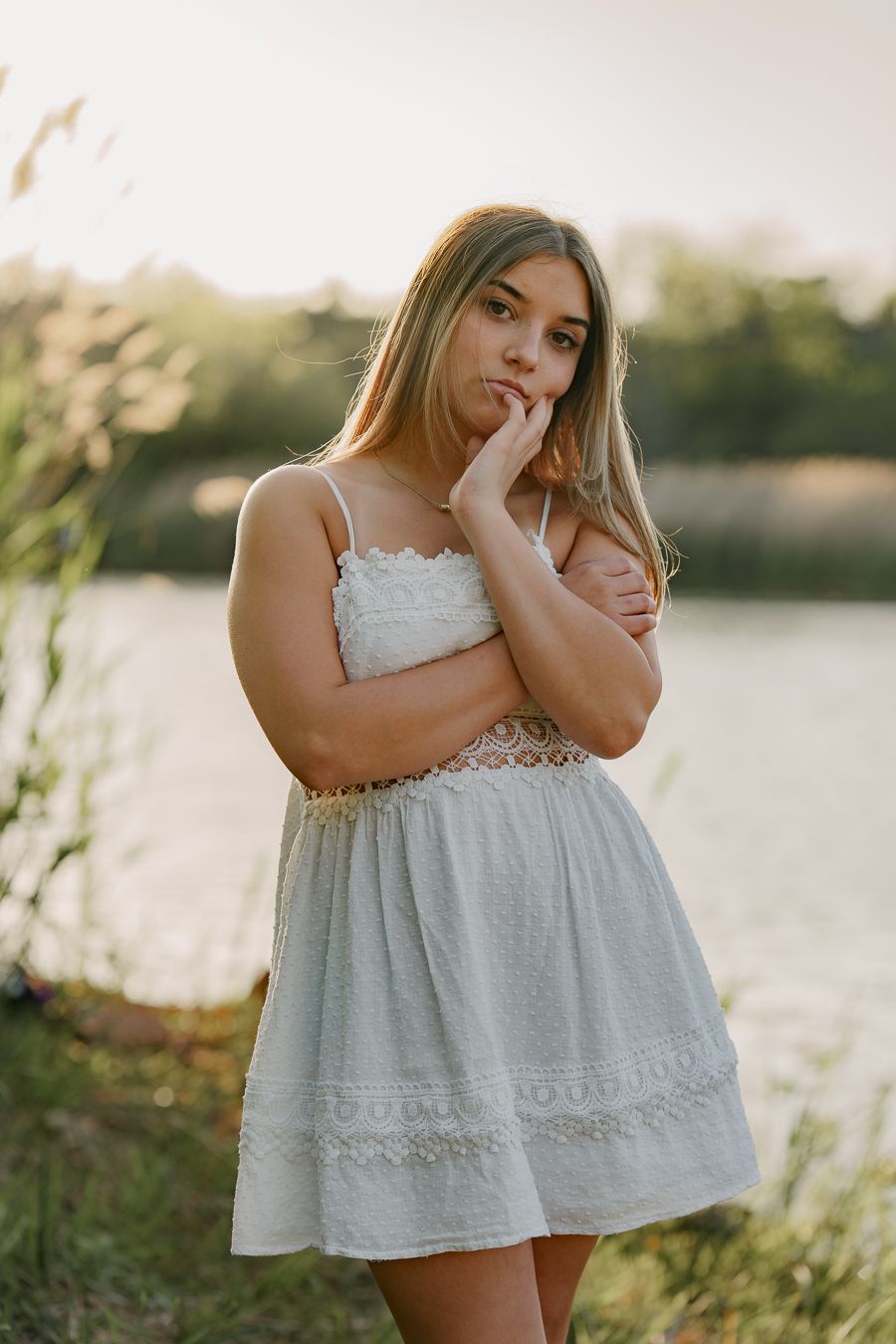 Young blonde woman in white lace sundress posing thoughtfully near a sunlit lake.